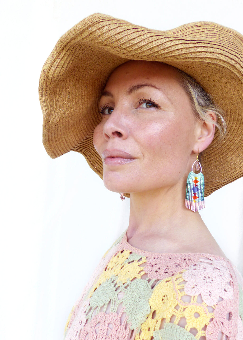 Peut inclure: Une femme porte un chapeau de paille à larges bords et des boucles d'oreilles en perles colorées. Les boucles d'oreilles présentent un motif rectangulaire avec des perles bleues, rouges et roses. Elle porte également un haut en crochet pastel.