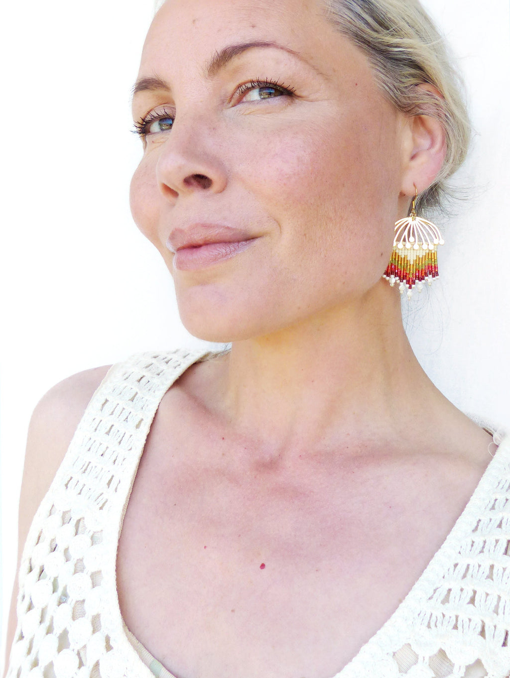 Peut inclure: Une femme portant un haut en crochet blanc avec une paire de boucles d'oreilles en perles dorées et multicolores. Les boucles d'oreilles sont en forme d'éventail et présentent des perles rouges, blanches et dorées.