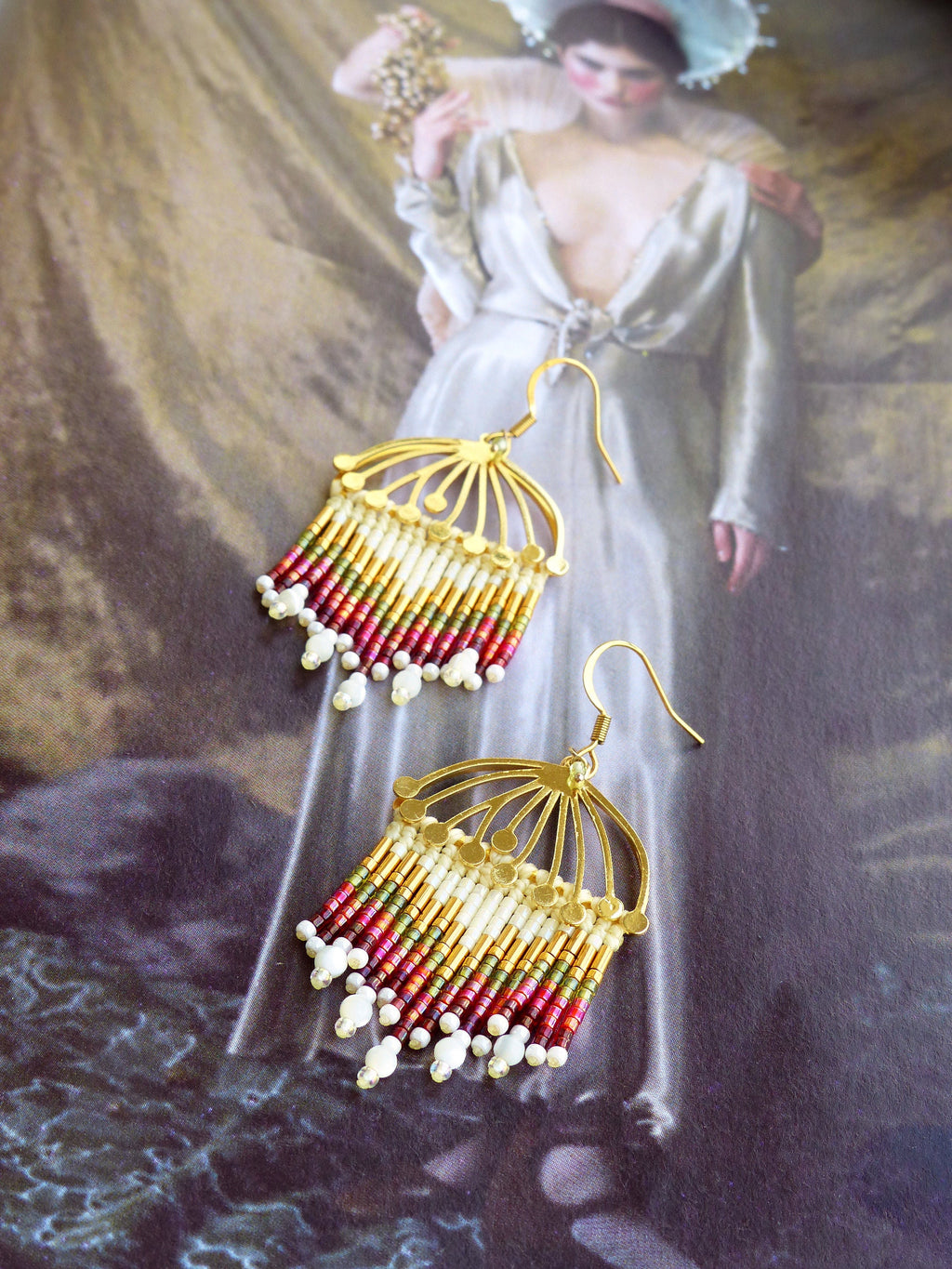 Peut inclure: Une paire de boucles d'oreilles en or avec un design délicat en forme d'éventail. Les boucles d'oreilles sont ornées de perles colorées dans des tons de rouge, vert et or, et présentent une frange de perles blanches.