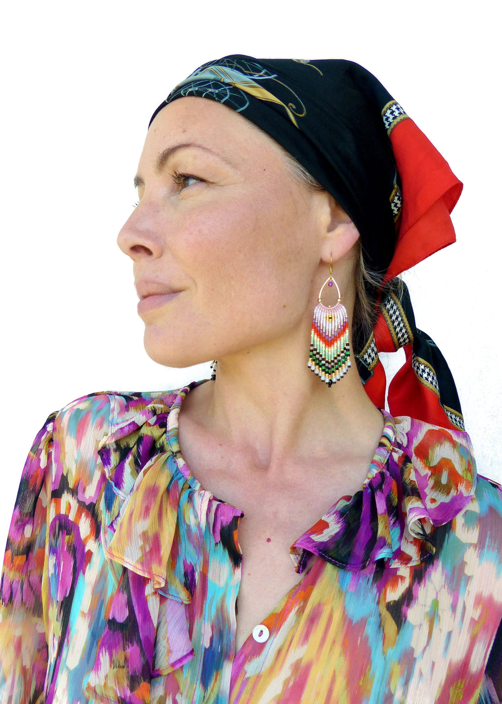 Peut inclure: Une femme portant un chemisier à motifs colorés avec un foulard noir et rouge. Elle porte de longues boucles d'oreilles en perles de couleur rose, blanche, verte et noire.