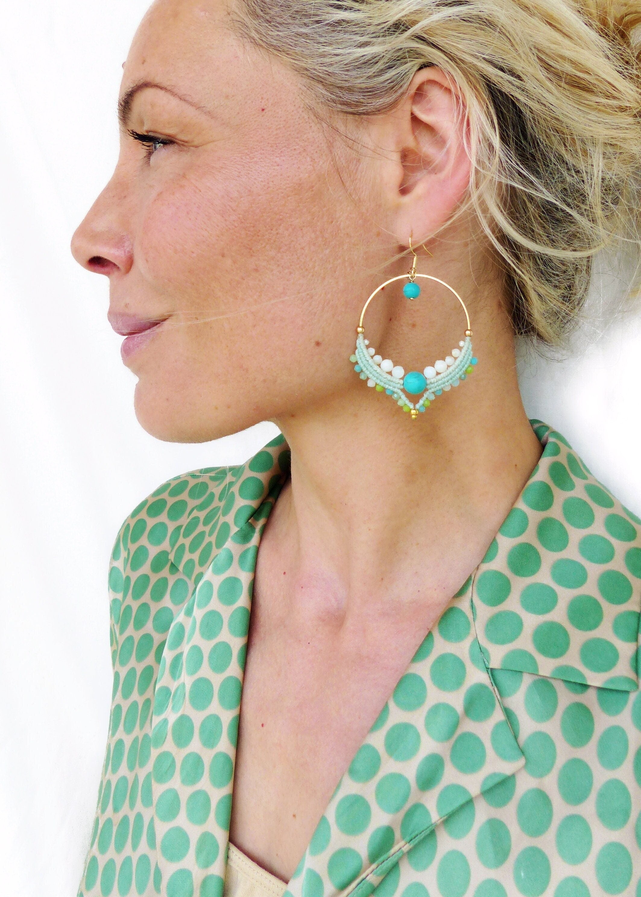 Peut inclure: Boucles d'oreilles créoles dorées avec des accents de perles turquoises et blanches. Les boucles d'oreilles présentent un design circulaire avec une perle turquoise centrale et de petites perles blanches, bleu clair et vertes. La femme porte une chemise à pois verts.
