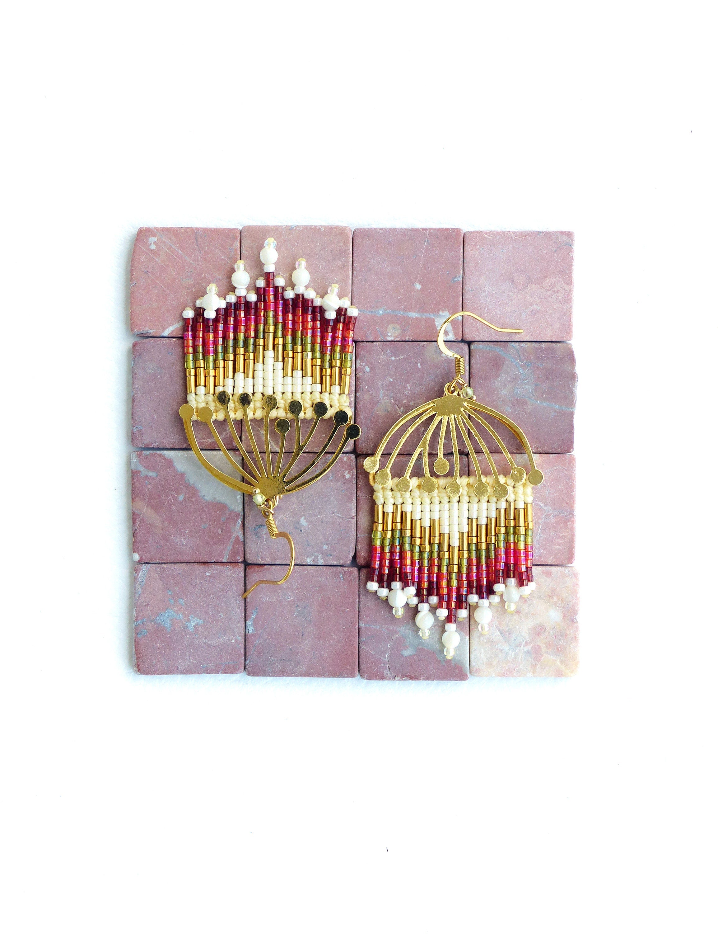 Peut inclure: Une paire de boucles d'oreilles créoles dorées avec des franges de perles rouges, dorées et blanches. Les boucles d'oreilles sont présentées sur une surface de carreaux roses.