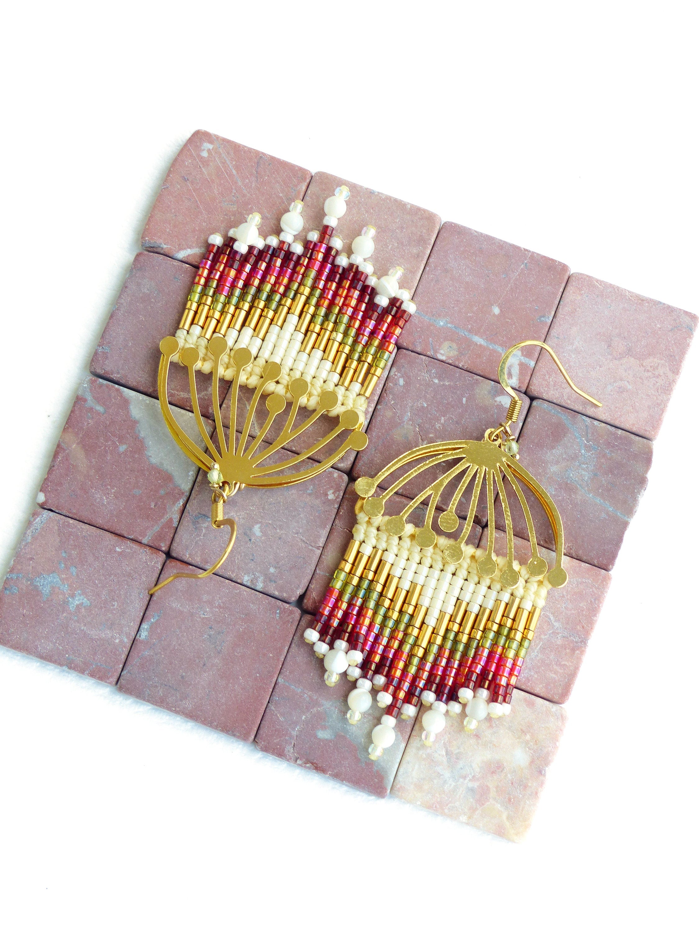 Peut inclure: Une paire de boucles d'oreilles en or avec une frange de perles. Les perles sont rouges, dorées et blanches. Les boucles d'oreilles sont suspendues à un fil d'or.
