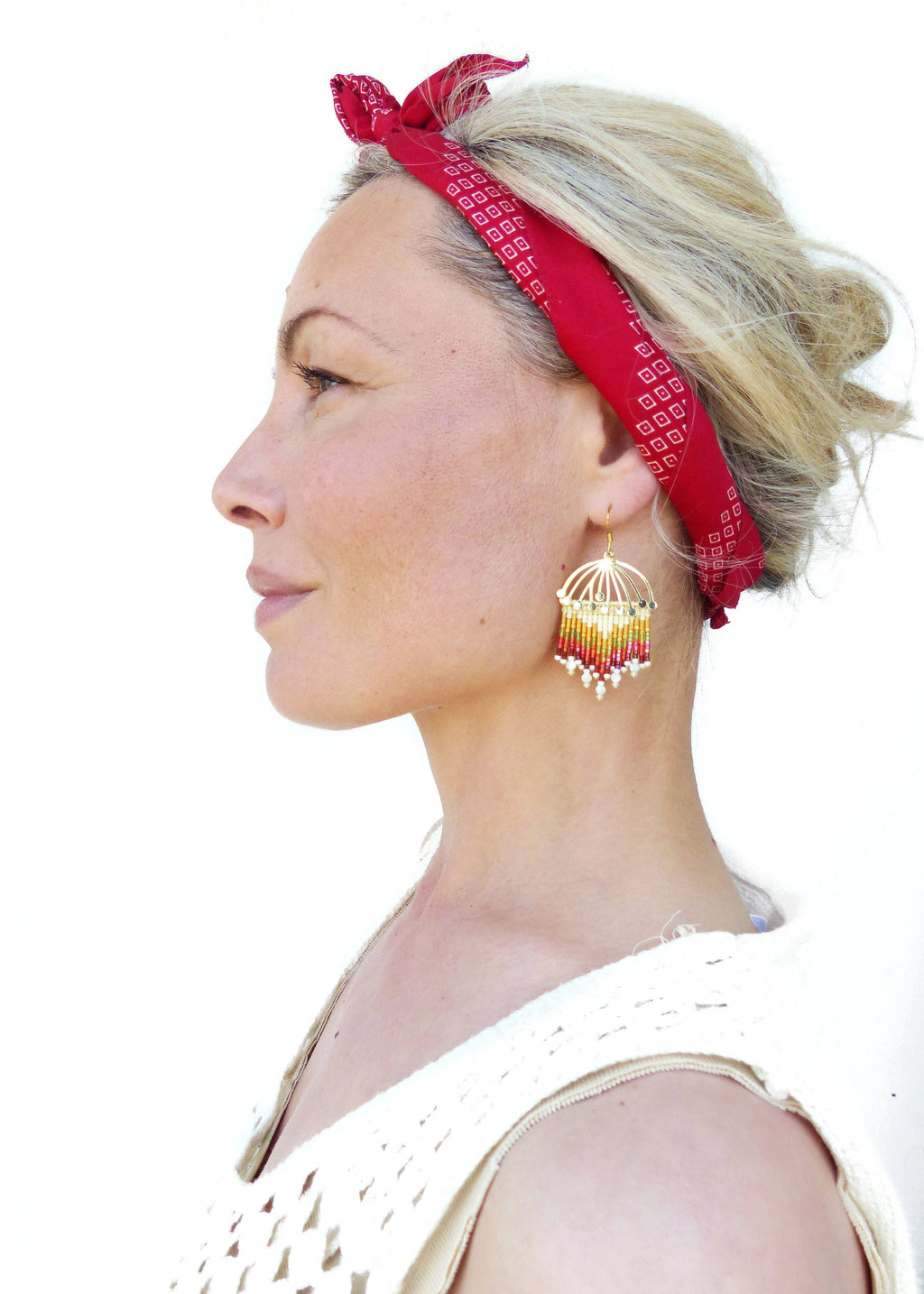 Peut inclure: Une femme aux cheveux blonds portant un bandana rouge avec un motif géométrique blanc et des boucles d'oreilles dorées avec des perles rouges, jaunes et blanches.