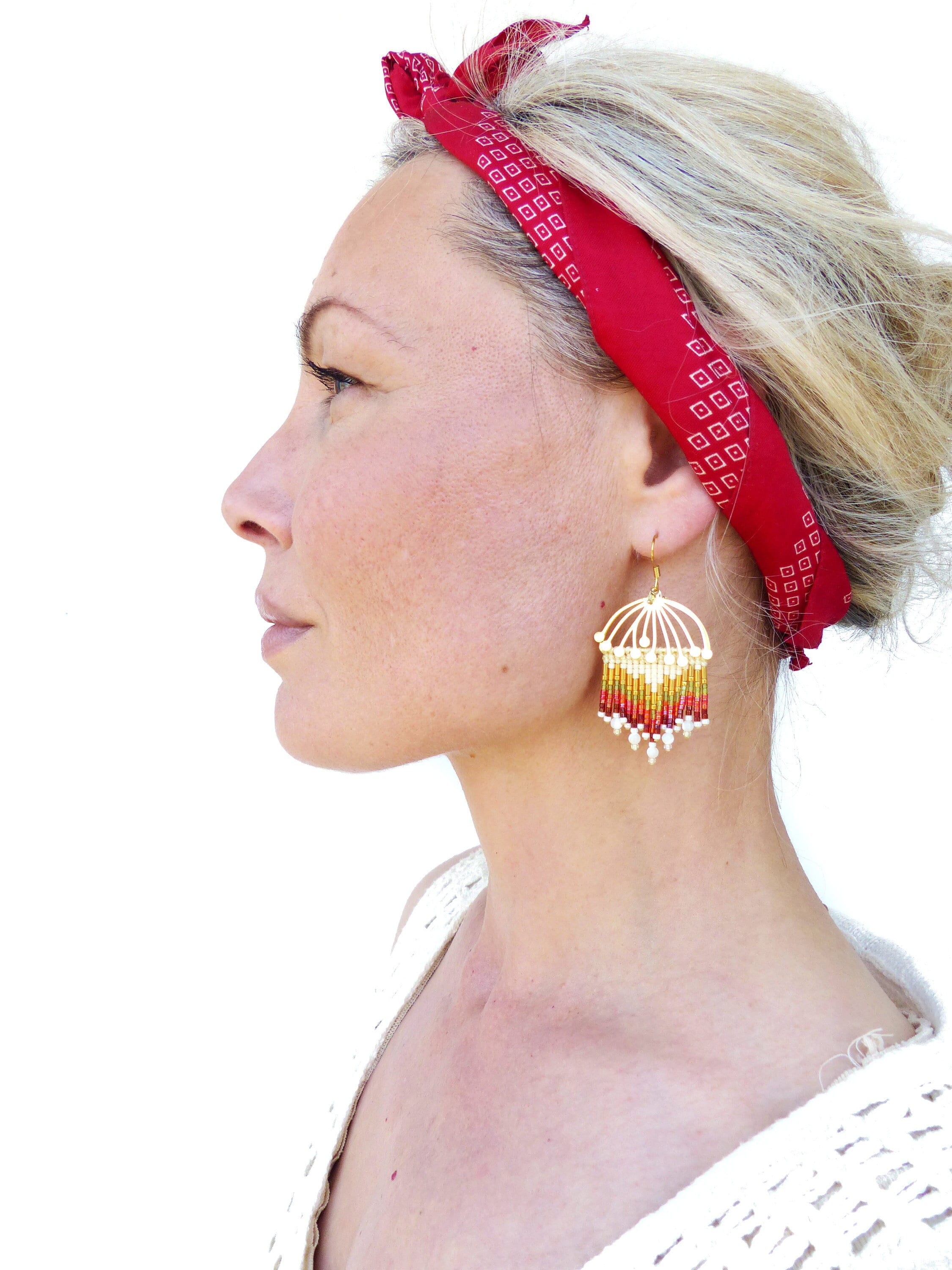 Peut inclure: Une femme portant un bandana rouge à motif géométrique et une paire de boucles d'oreilles en perles dorées et multicolores. Les boucles d'oreilles sont longues et pendantes, avec une forme de larme.