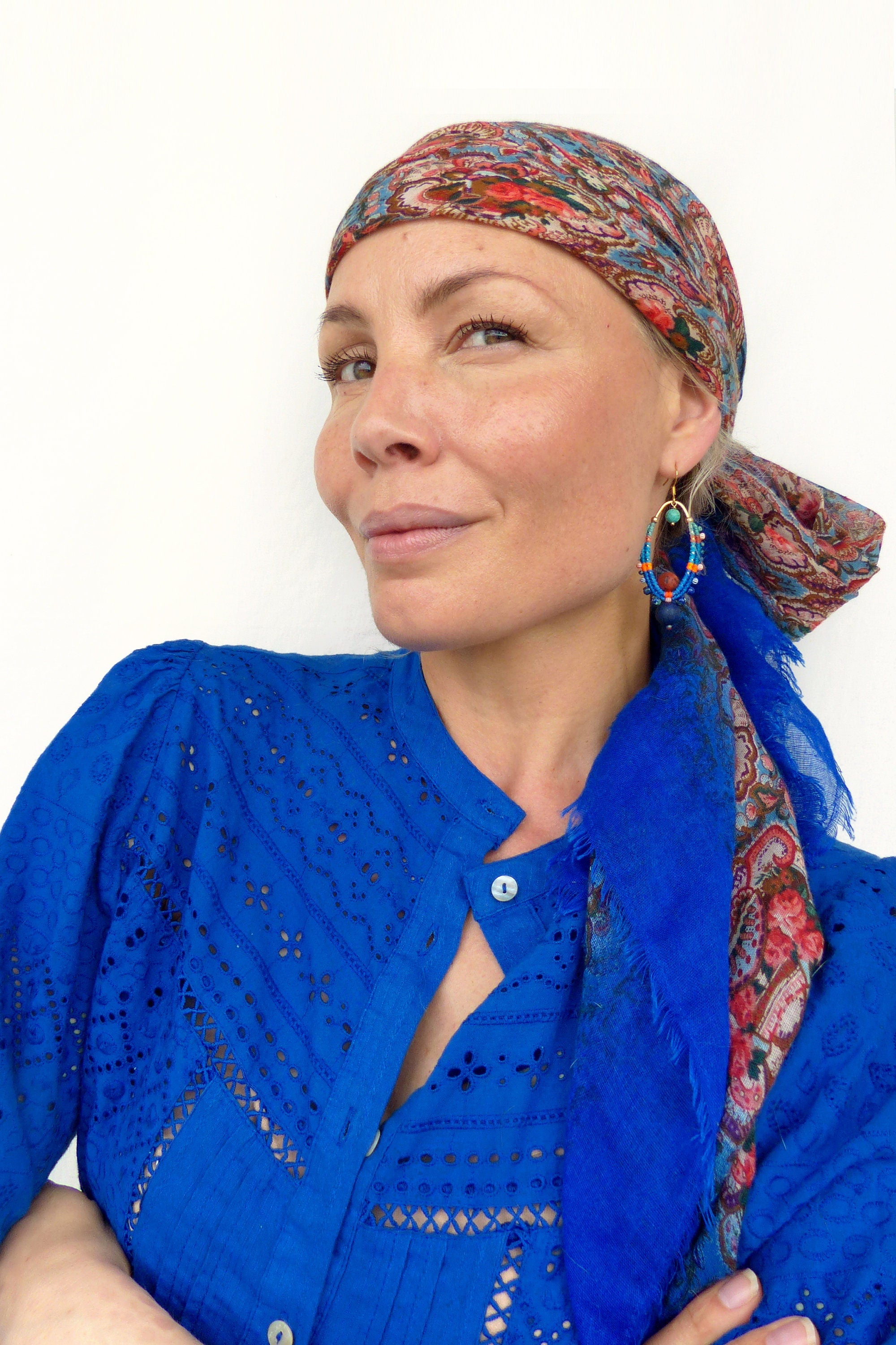 Peut inclure: Une femme portant une chemise bleue brodée avec un foulard paisley coloré noué autour de sa tête. Elle porte de grandes boucles d'oreilles en forme de cerceau avec des perles colorées.