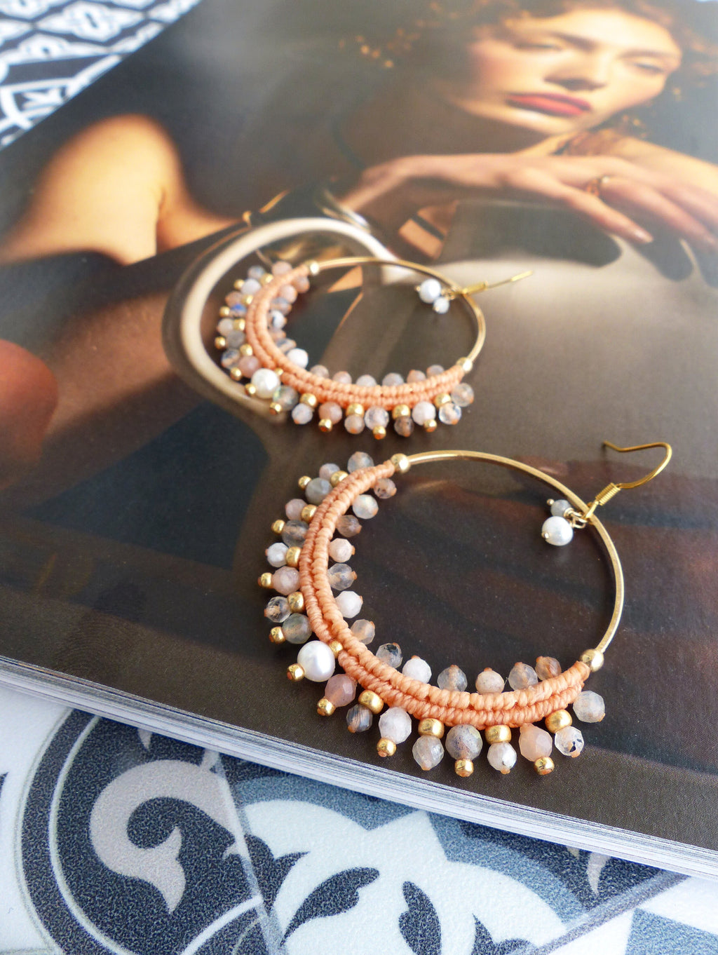Peut inclure: Une paire de boucles d'oreilles créoles dorées avec des perles de couleur pêche et des perles blanches. Les boucles d'oreilles sont sur un magazine avec un visage de femme sur la couverture.