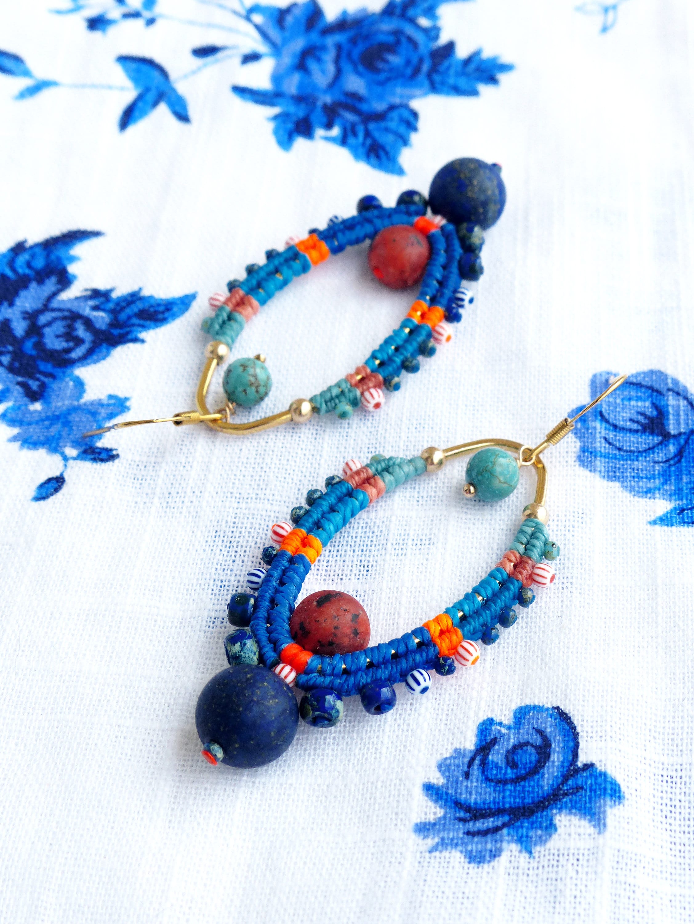 Peut inclure: Une paire de boucles d'oreilles en perles bleues, orange et rouges avec une pierre de lapis-lazuli sur chaque boucle d'oreille. Les boucles d'oreilles sont suspendues à des créoles dorées.
