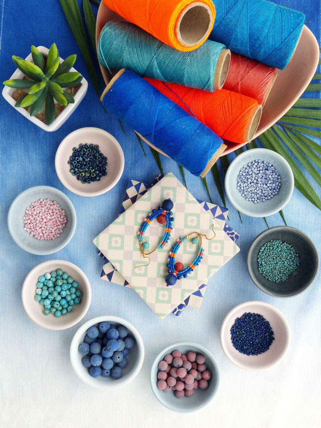 Peut inclure: Assortiment de perles colorées dans de petits bols, y compris des perles turquoise, roses, bleues et brunes. Une paire de boucles d'oreilles faites à la main avec des perles bleues, rouges et oranges est présentée sur un papier à motifs.