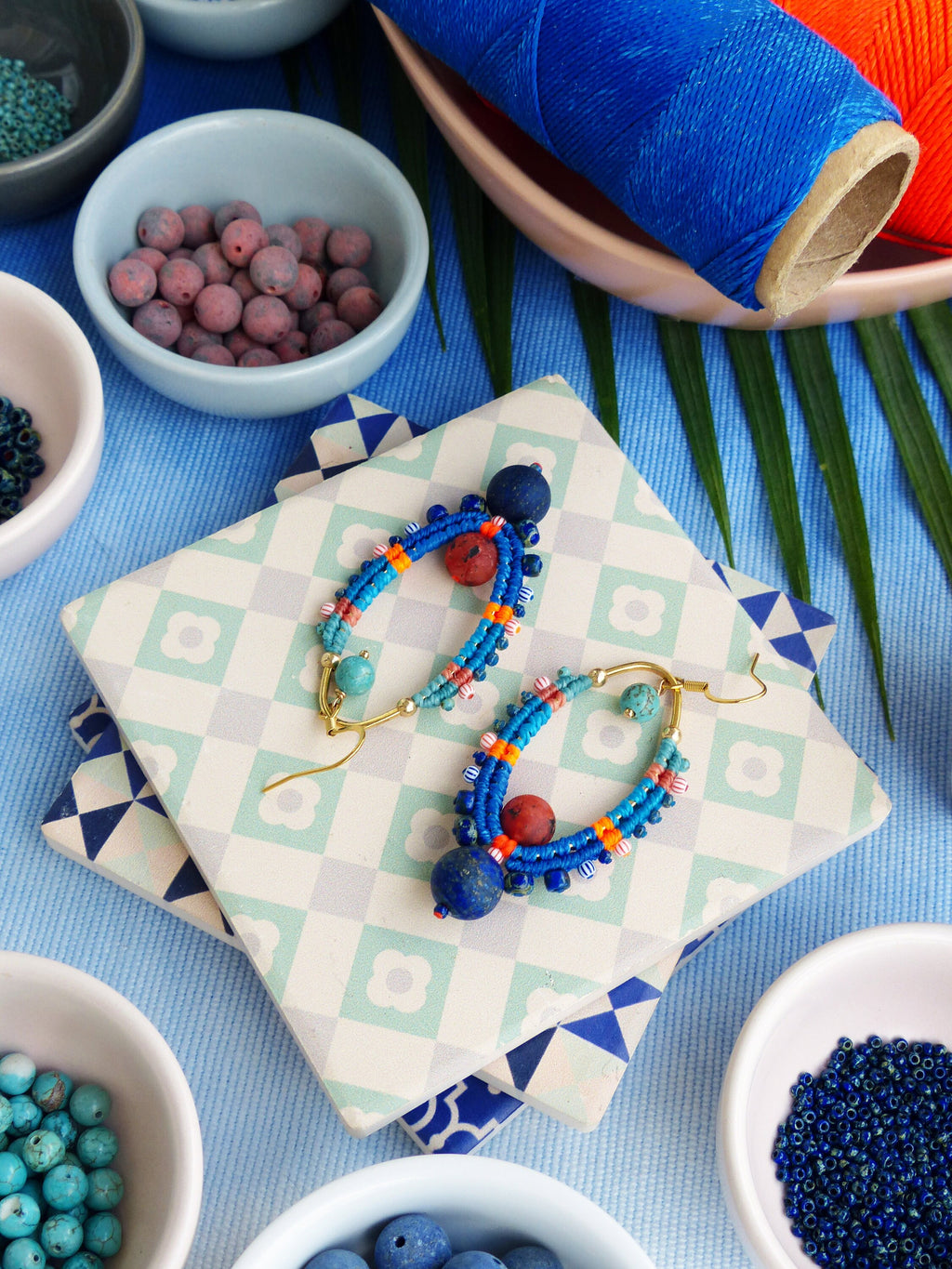 Peut inclure: Deux paires de boucles d'oreilles faites à la main avec des perles bleues, orange et rouges. Les boucles d'oreilles sont sur une surface carrelée blanche et bleue.