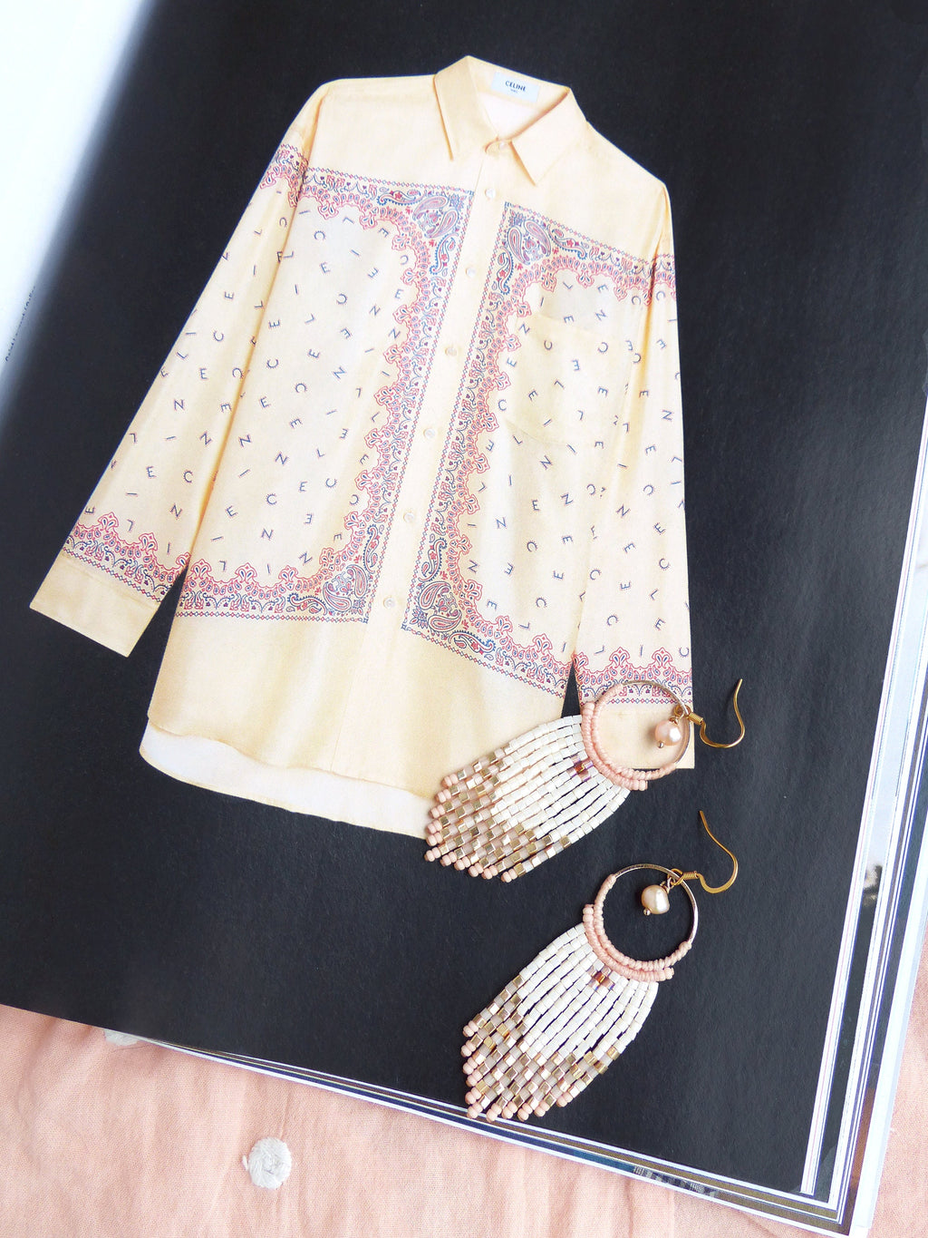 Peut inclure: Une paire de longues boucles d'oreilles en perles avec un anneau en or et un accent de perle. Les boucles d'oreilles sont blanches et roses avec un design en couches. Les boucles d'oreilles sont photographiées sur un fond noir avec une chemise jaune à motif paisley.