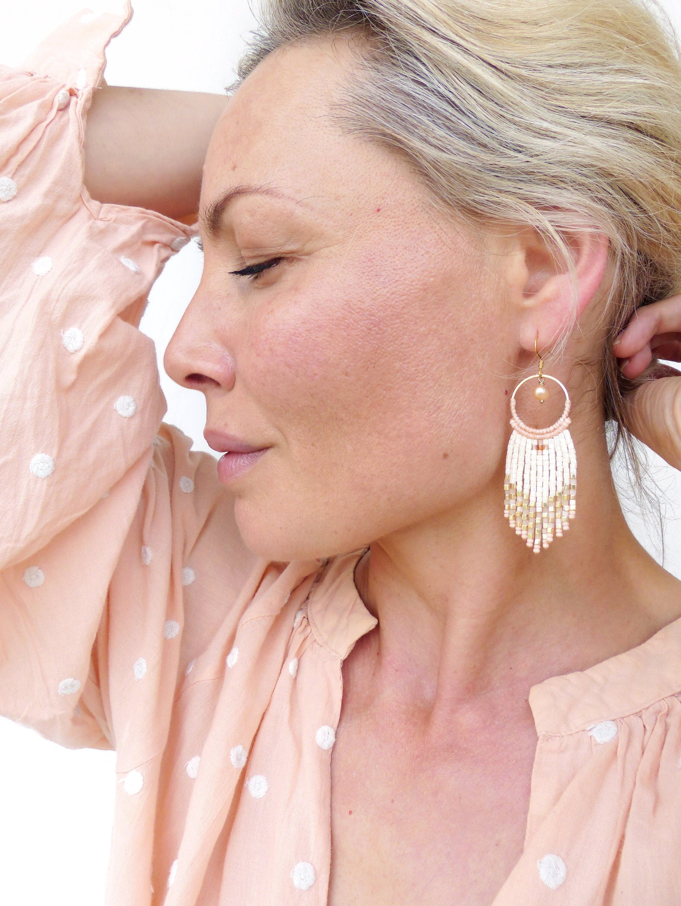 Peut inclure: Une paire de boucles d'oreilles en perles blanches et pêche avec un anneau doré et une perle dorée en haut. Les boucles d'oreilles pendent de l'oreille d'une personne aux cheveux blonds.