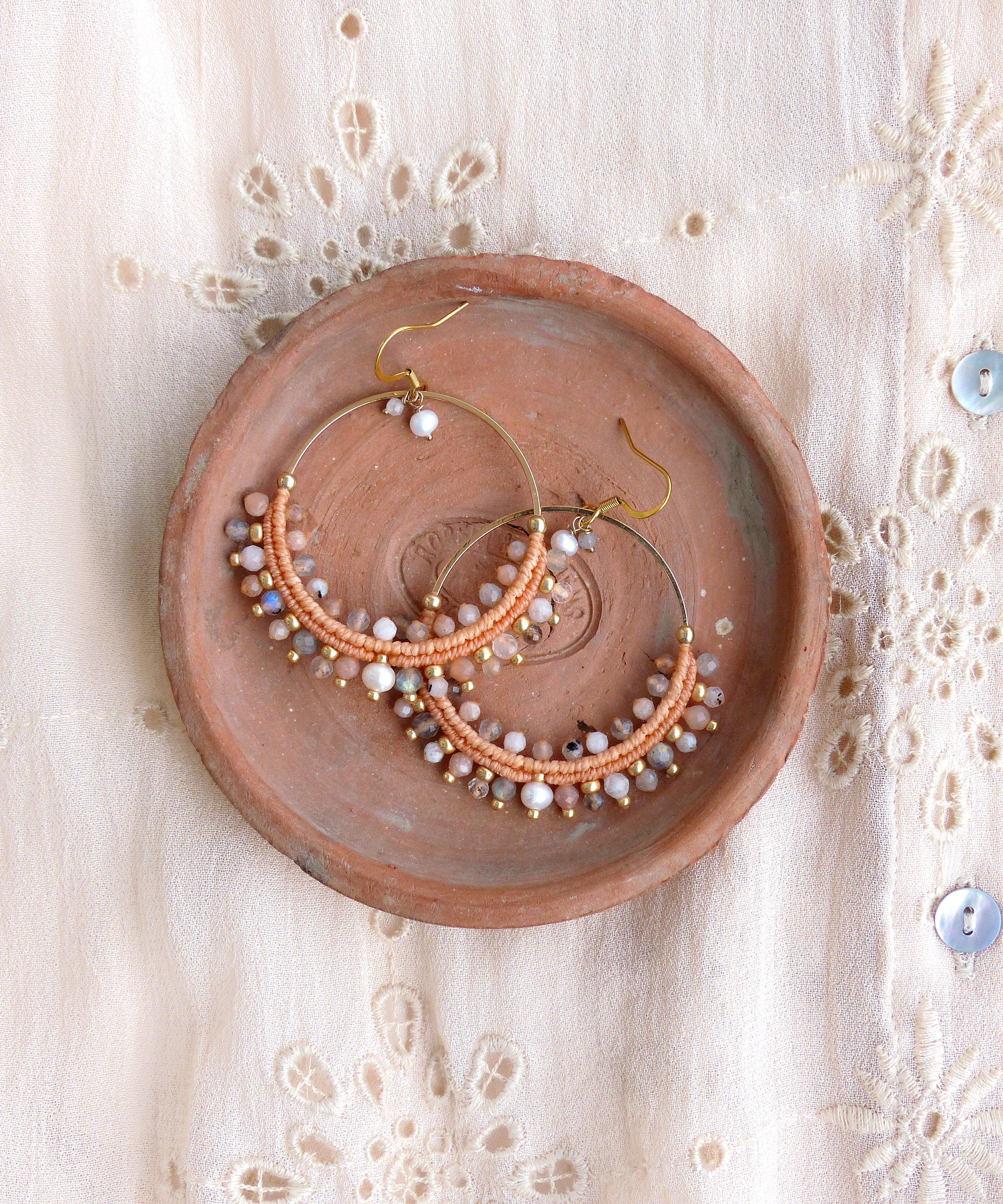 Peut inclure: Une paire de boucles d'oreilles créoles de couleur pêche avec un fil d'or et une variété de petites perles irisées. Les boucles d'oreilles sont présentées sur une assiette en terre cuite.