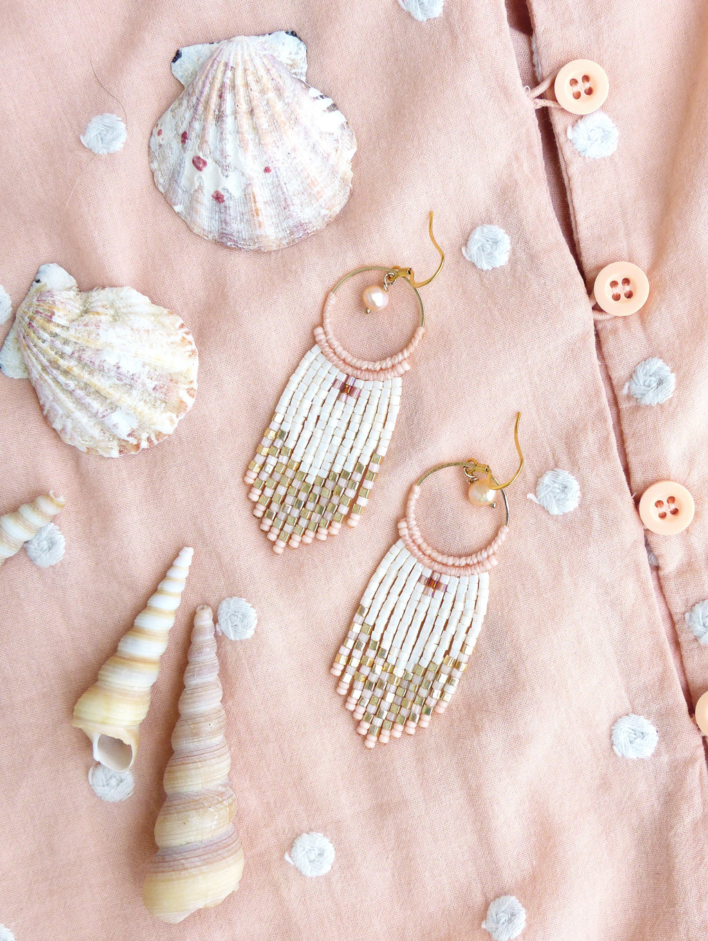Peut inclure: Une paire de boucles d'oreilles créoles dorées avec des franges de perles blanches et roses. Les boucles d'oreilles sont sur un fond de tissu rose à pois blancs et des coquillages.