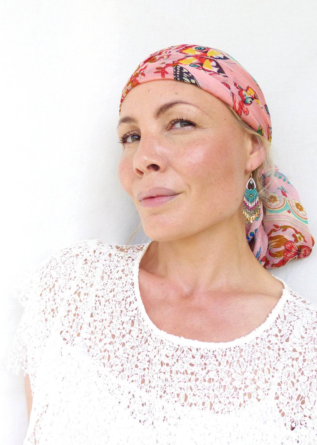 Peut inclure: Une femme aux cheveux châtains clairs porte un foulard rose à motifs floraux et un haut en dentelle blanche. Elle porte des boucles d'oreilles en perles turquoise et dorées.
