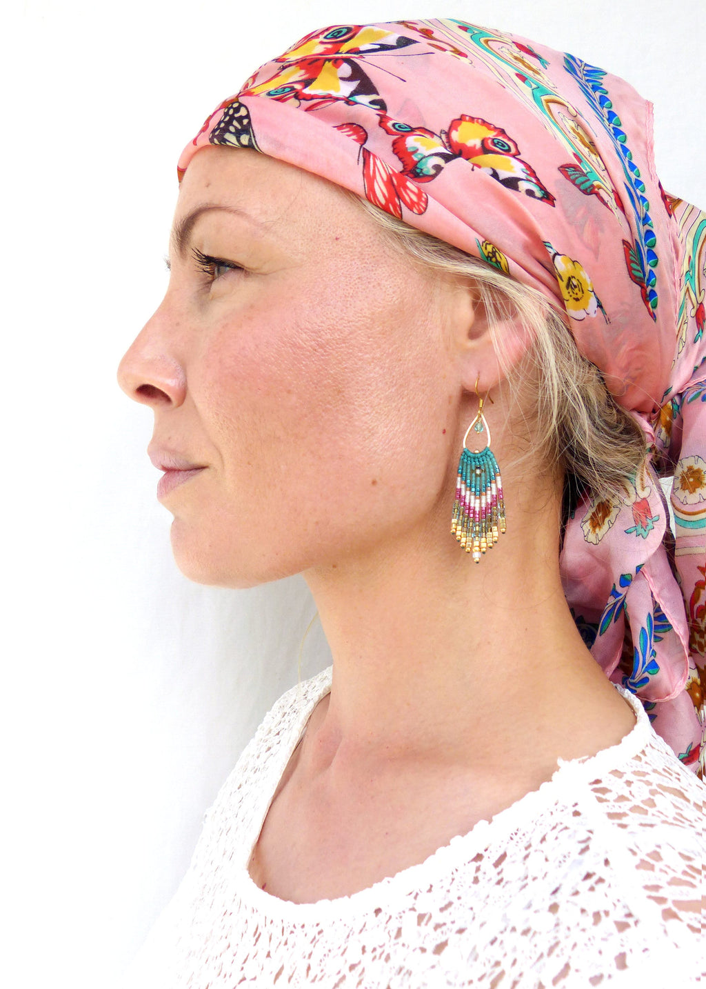 Peut inclure: Une femme portant un foulard rose à imprimé papillon et un haut en crochet blanc. Elle porte de longues boucles d'oreilles en perles turquoise, dorées et blanches.