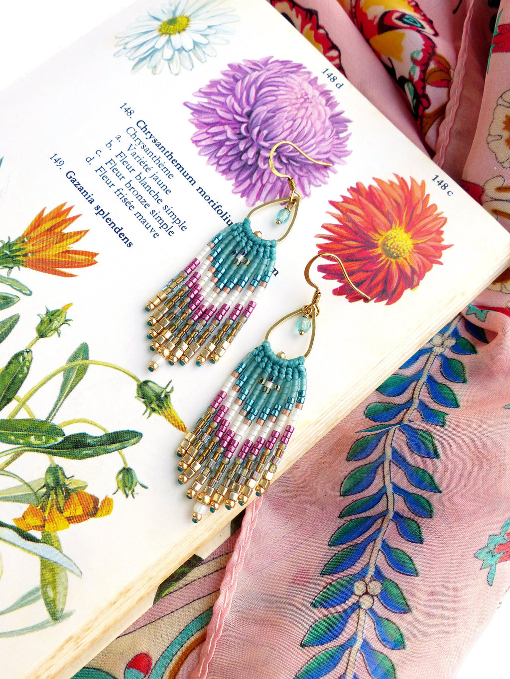 Peut inclure: Une paire de boucles d'oreilles en perles turquoise, blanches, dorées et roses avec des crochets dorés. Les boucles d'oreilles sont suspendues à un livre avec des illustrations botaniques et un tissu rose à motif floral.