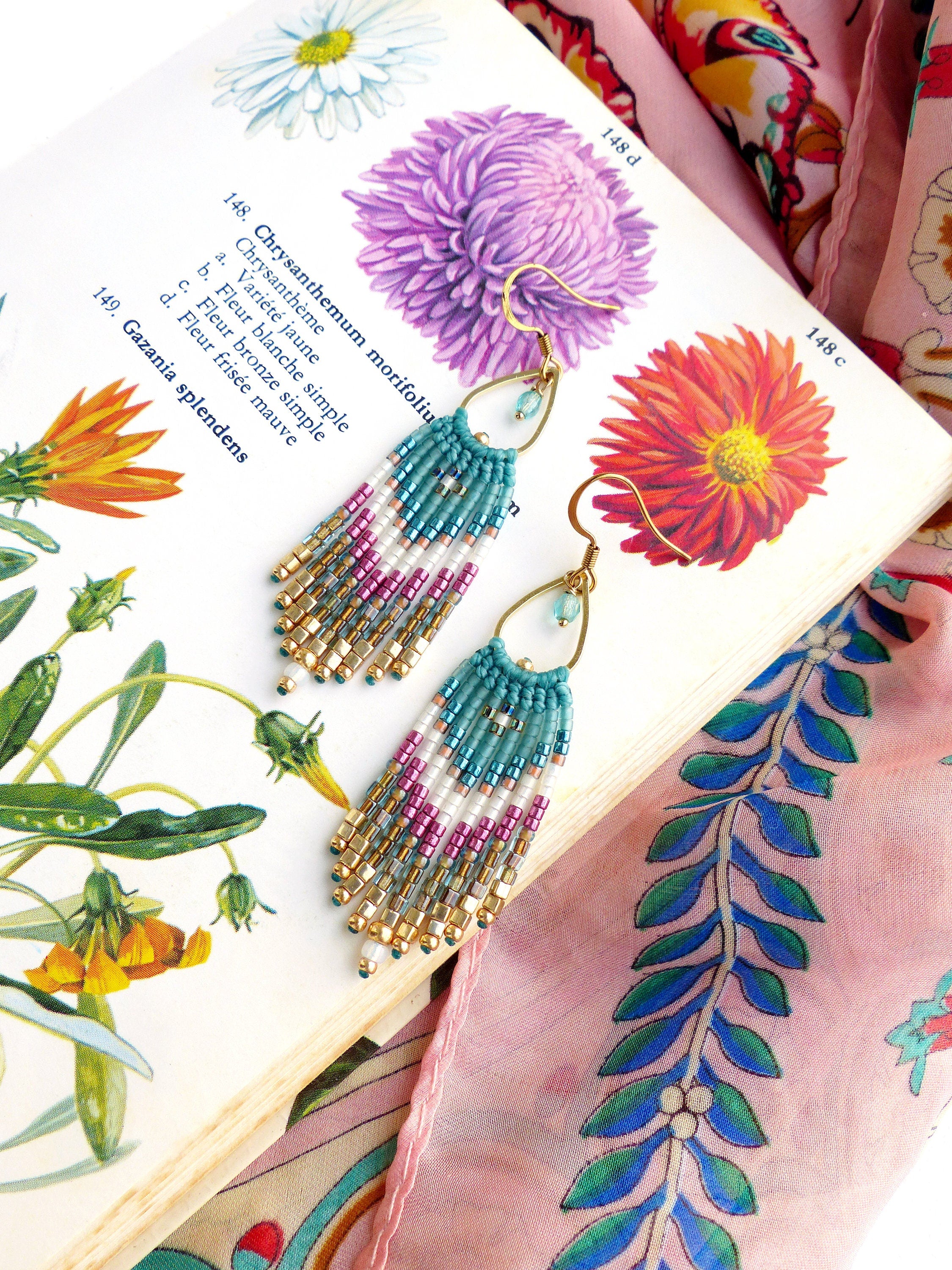 Peut inclure: Une paire de boucles d'oreilles en perles turquoise, blanches, dorées et roses avec des crochets dorés. Les boucles d'oreilles sont suspendues à un livre avec des illustrations botaniques et un tissu rose à motif floral.