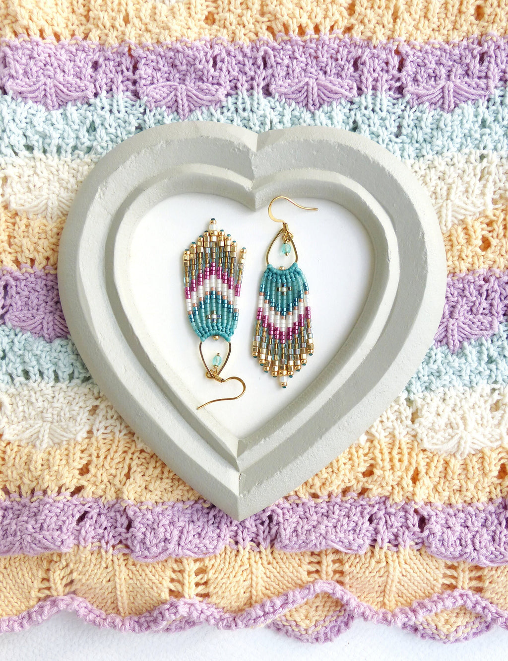 Peut inclure: Une paire de boucles d'oreilles pendantes avec un motif en chevron composé de perles turquoise, blanches et dorées. Les boucles d'oreilles sont présentées sur un plateau en forme de cœur.