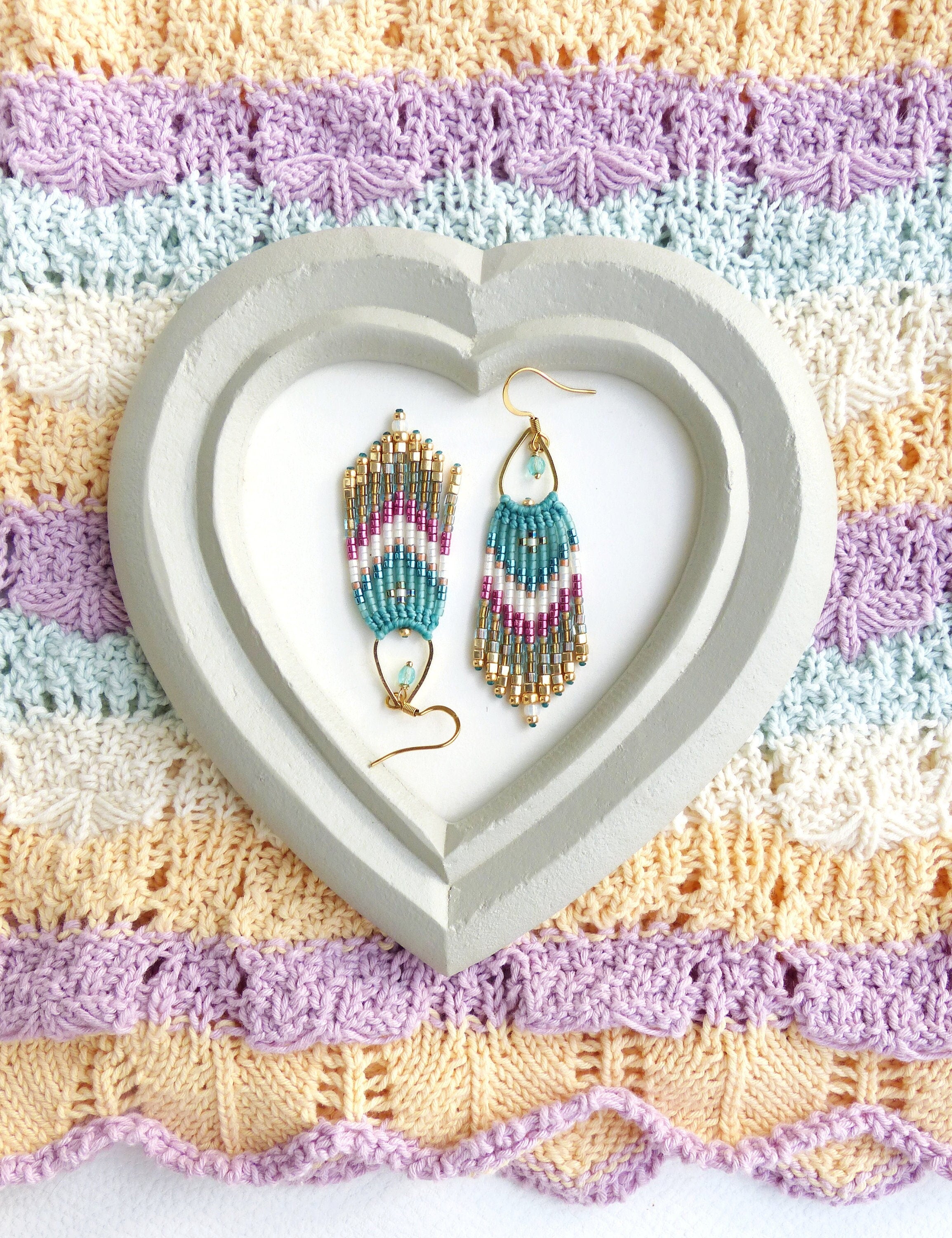 Peut inclure: Une paire de boucles d'oreilles pendantes avec un motif en chevron composé de perles turquoise, blanches et dorées. Les boucles d'oreilles sont présentées sur un plateau en forme de cœur.