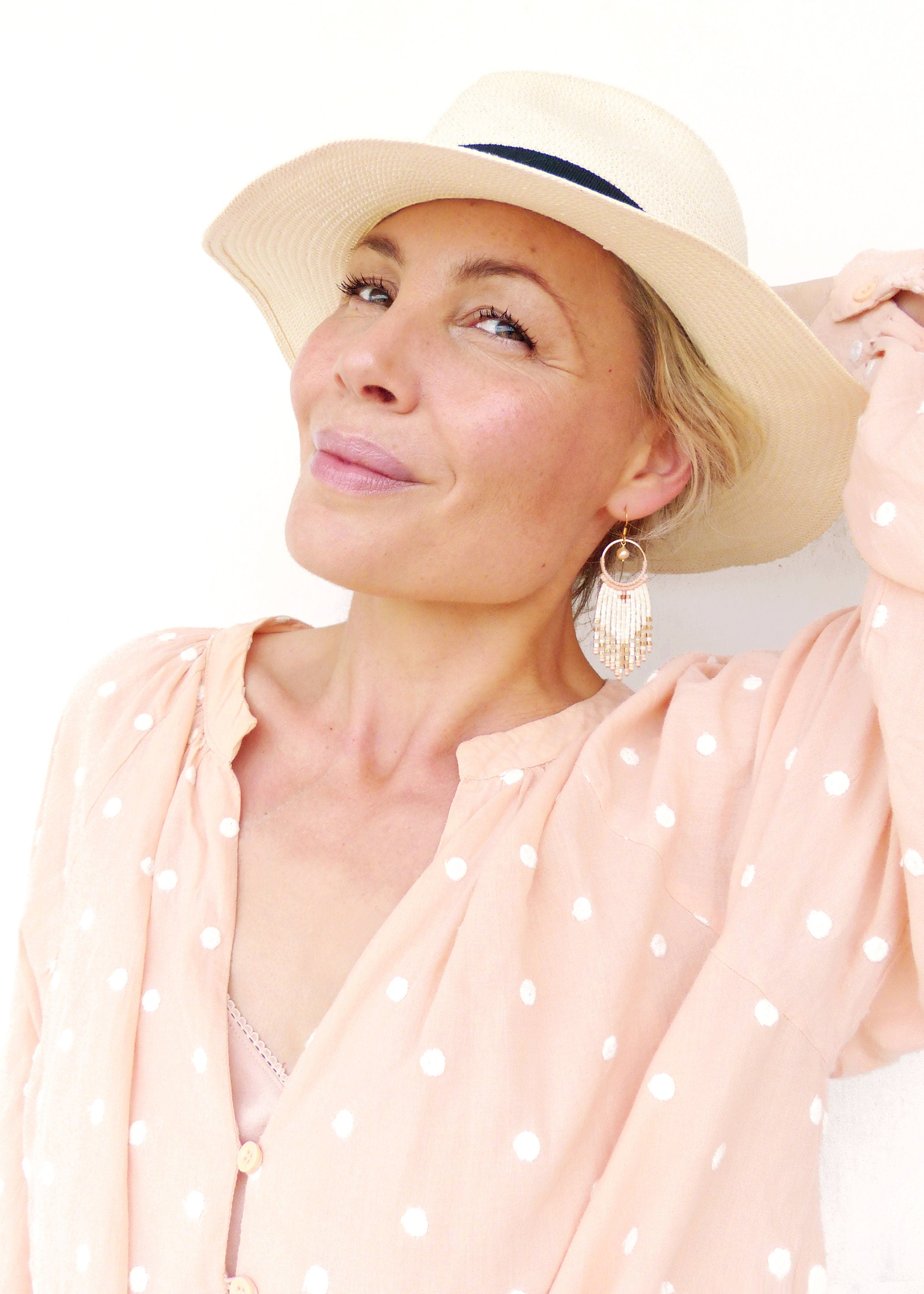 Peut inclure: Une femme portant un chapeau de paille blanc avec un bandeau noir, une blouse à pois rose pêche et des boucles d'oreilles en perles blanches. 