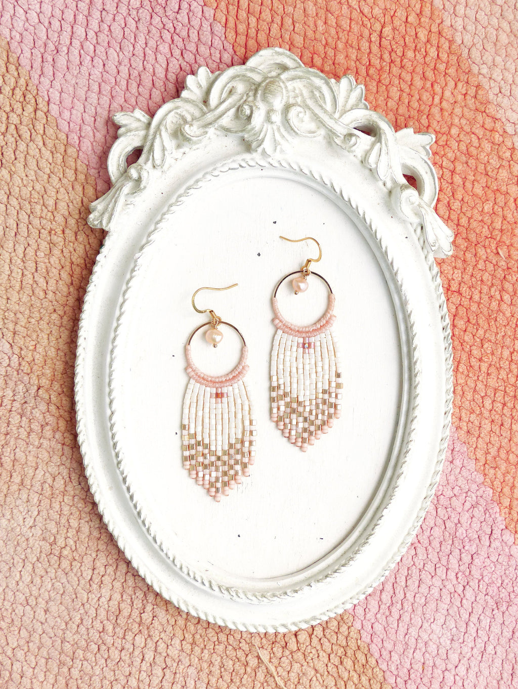 Peut inclure: Une paire de boucles d'oreilles créoles dorées avec des franges de perles roses et blanches. Les boucles d'oreilles sont présentées sur un cadre ovale blanc.