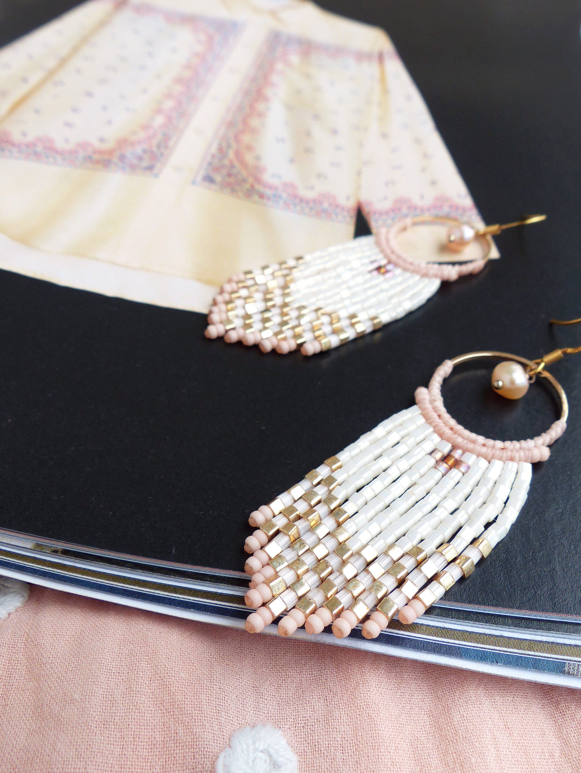 Peut inclure: Une paire de boucles d'oreilles en franges de perles blanches et dorées avec des accents roses. Les boucles d'oreilles sont suspendues à un magazine.