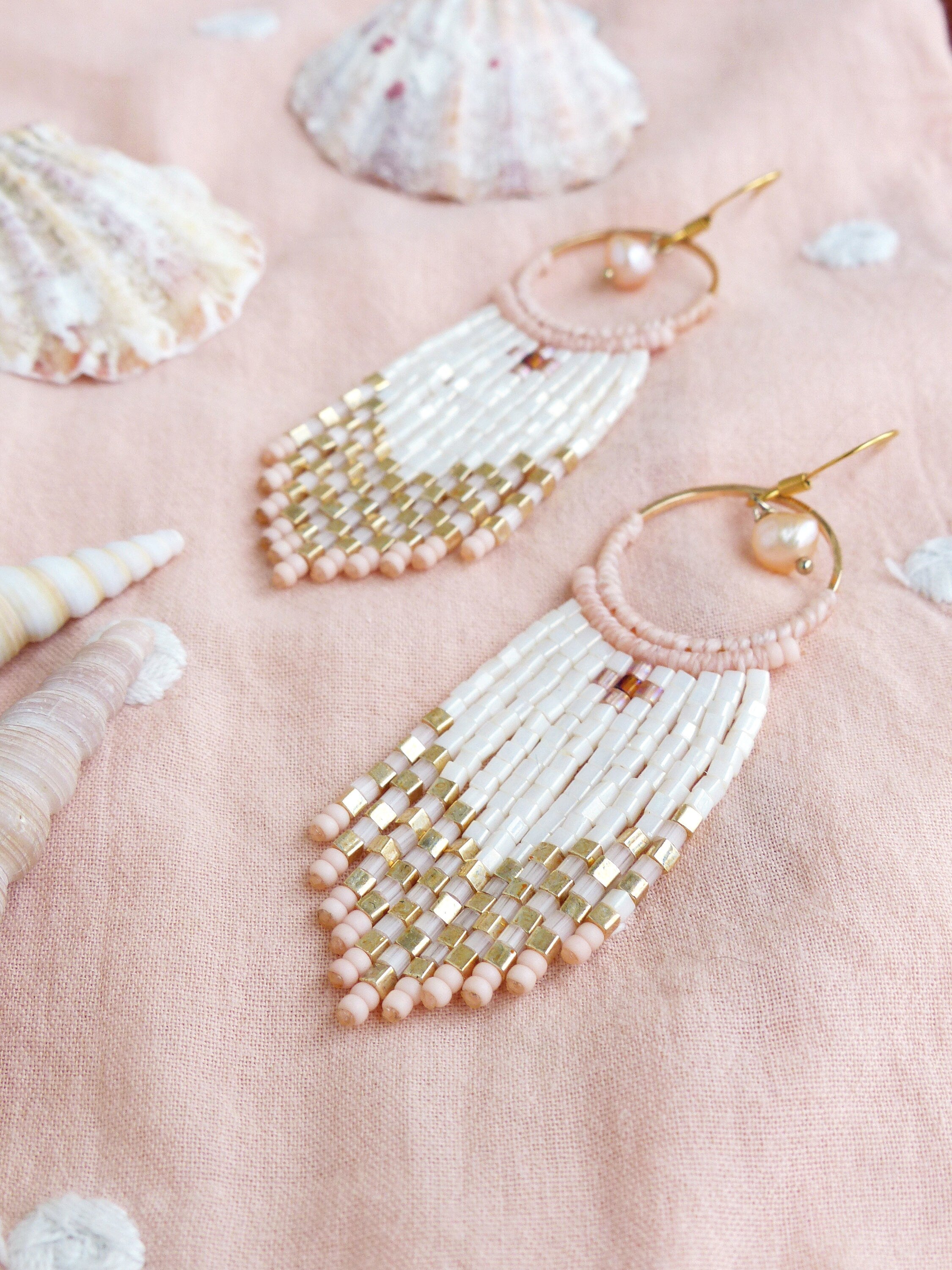 Peut inclure: Une paire de longues boucles d'oreilles en perles avec un schéma de couleurs rose et blanc. Les boucles d'oreilles présentent un anneau en or avec un petit charme en perle et une frange de perles roses et blanches.