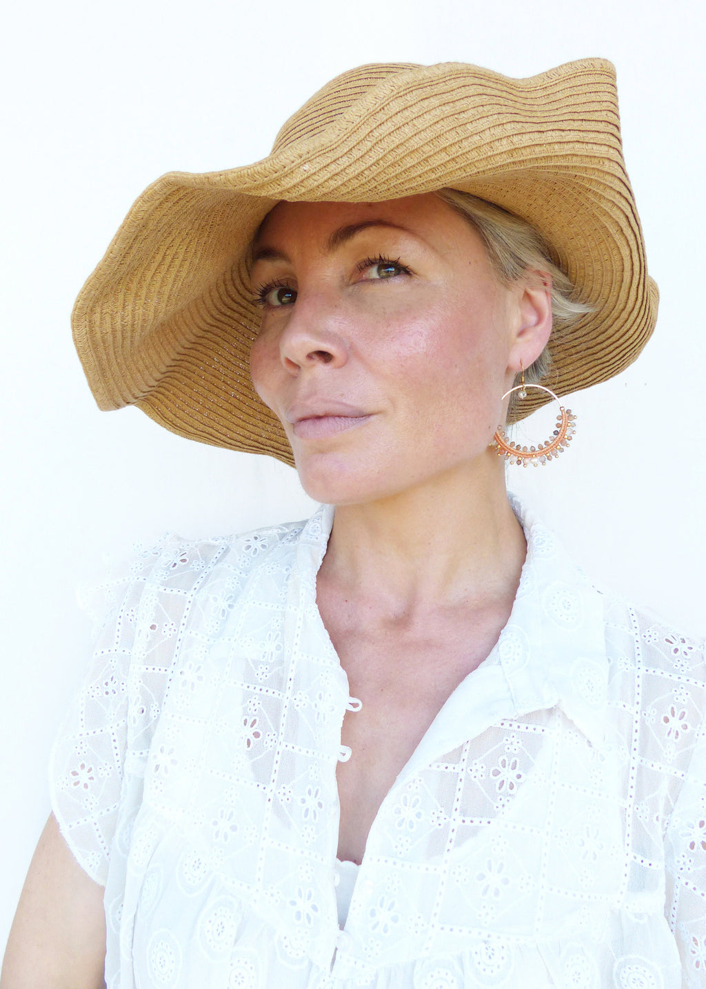 Peut inclure: Une femme portant un chapeau de paille à larges bords et une blouse blanche en broderie anglaise avec un motif floral délicat. Elle porte de grandes boucles d'oreilles créoles avec de petites perles colorées.