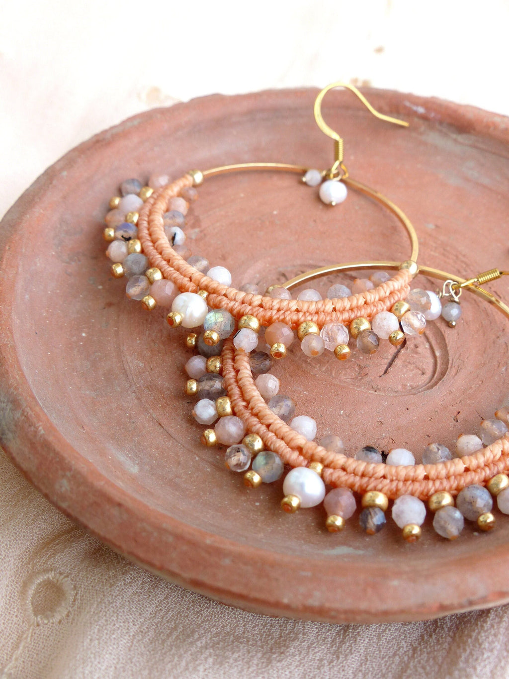 Peut inclure: Une paire de boucles d'oreilles créoles dorées avec un cordon de couleur pêche enroulé autour du cercle et rehaussé de petites pierres claires et de perles blanches.