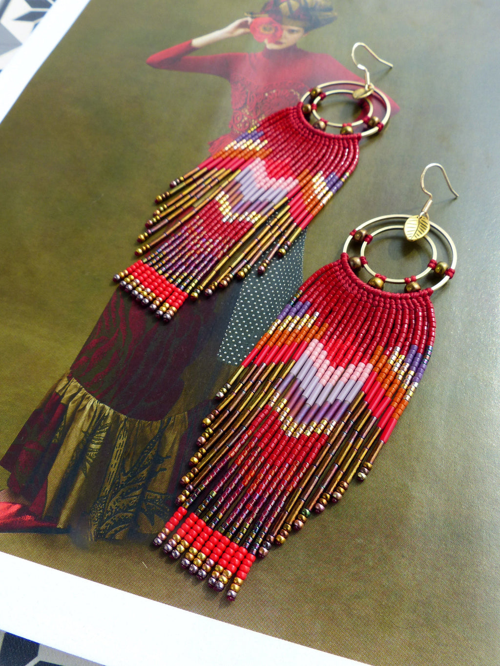 Peut inclure: Une paire de longues boucles d'oreilles rouges en perles avec des cercles dorés et un charme en forme de feuille. Les boucles d'oreilles présentent un motif géométrique avec des perles rouges, orange, violettes et dorées.
