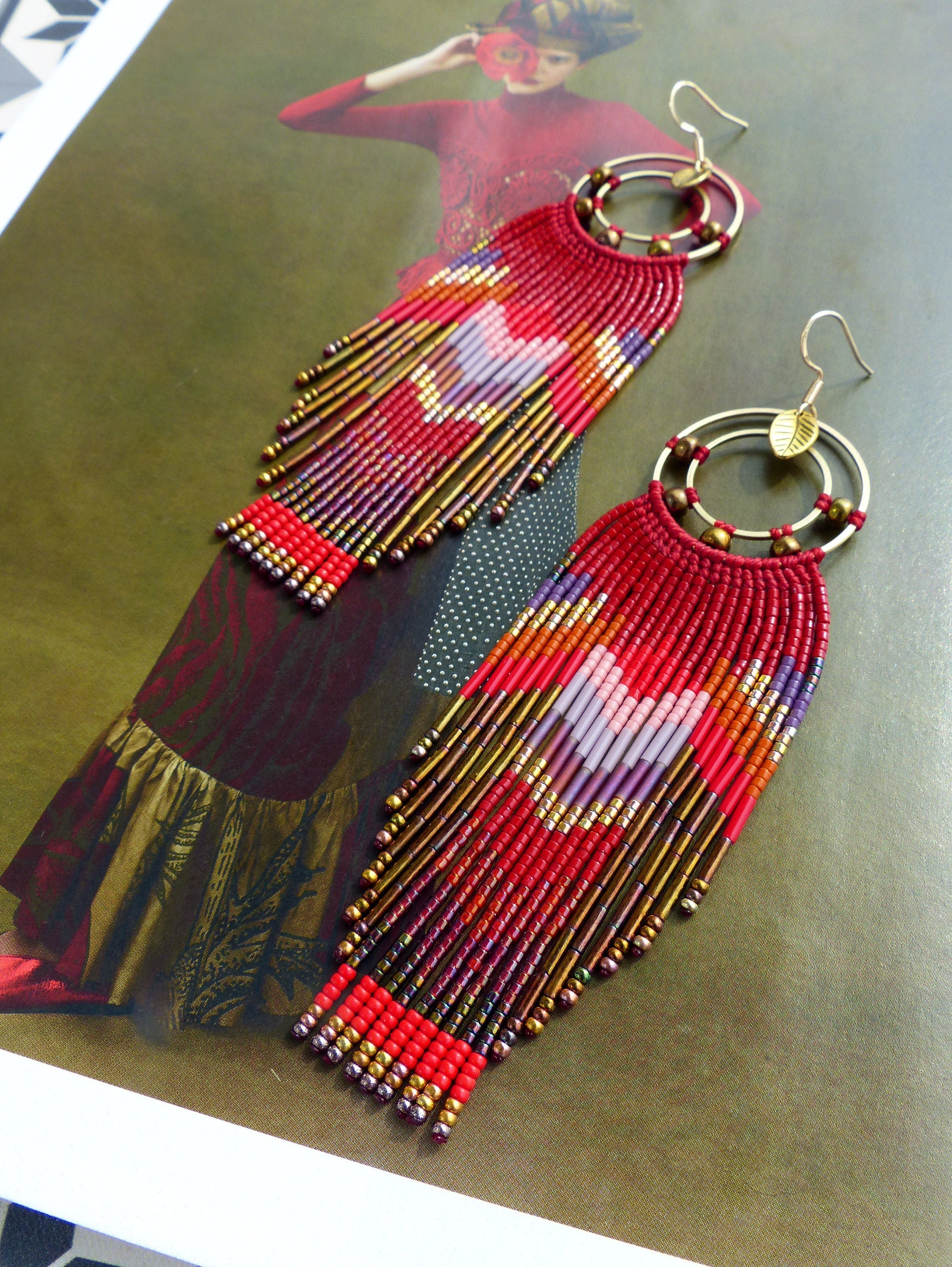 Peut inclure: Une paire de longues boucles d'oreilles rouges en perles avec des cercles dorés et un charme en forme de feuille. Les boucles d'oreilles présentent un motif géométrique avec des perles rouges, orange, violettes et dorées.