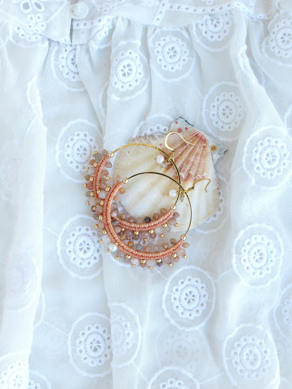 Peut inclure: Une paire de boucles d'oreilles créoles dorées avec des perles roses et brunes. Les boucles d'oreilles sont sur un fond de tissu en dentelle blanche.