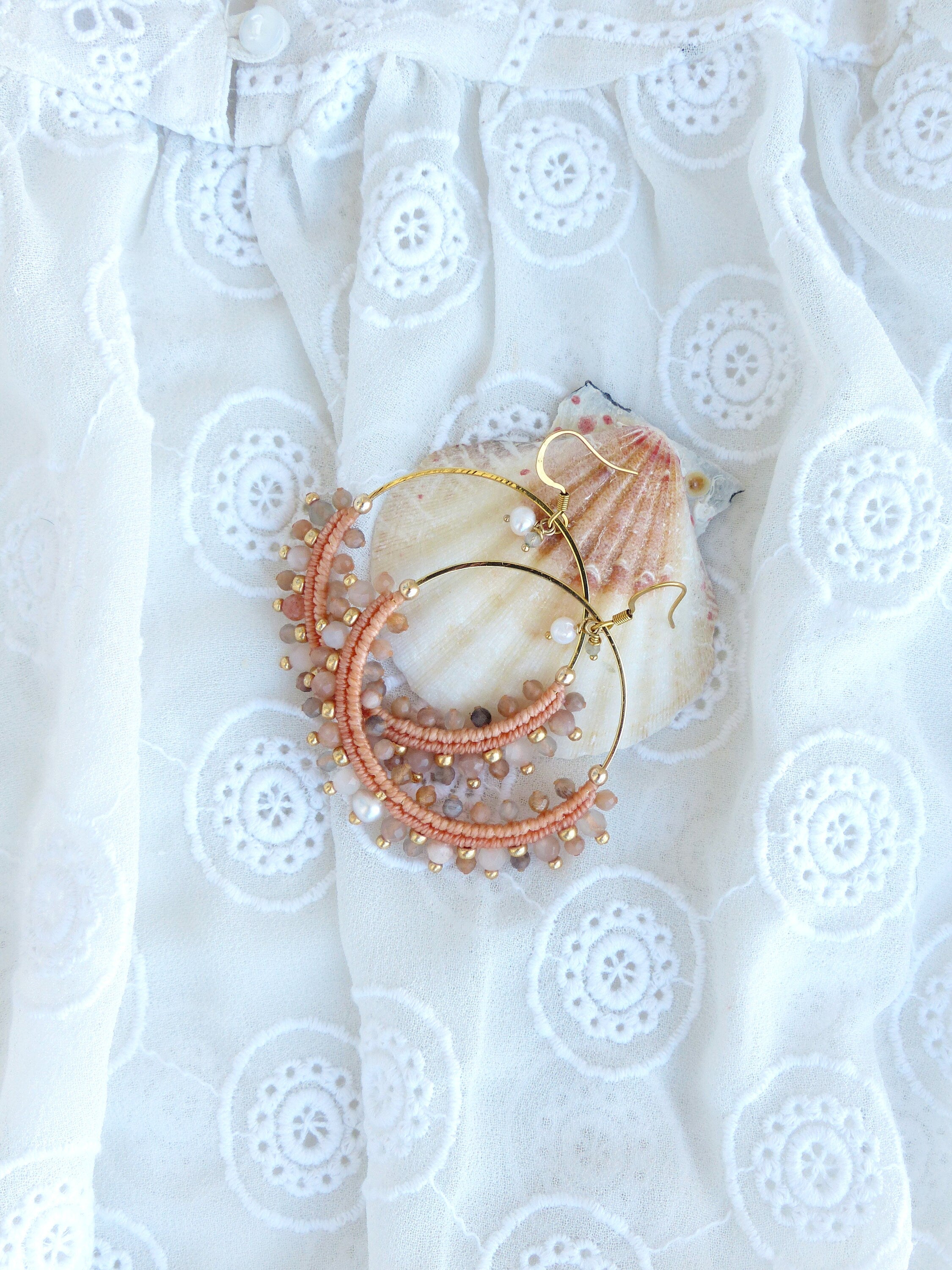 Peut inclure: Une paire de boucles d'oreilles créoles dorées avec des perles roses et brunes. Les boucles d'oreilles sont sur un fond de tissu en dentelle blanche.