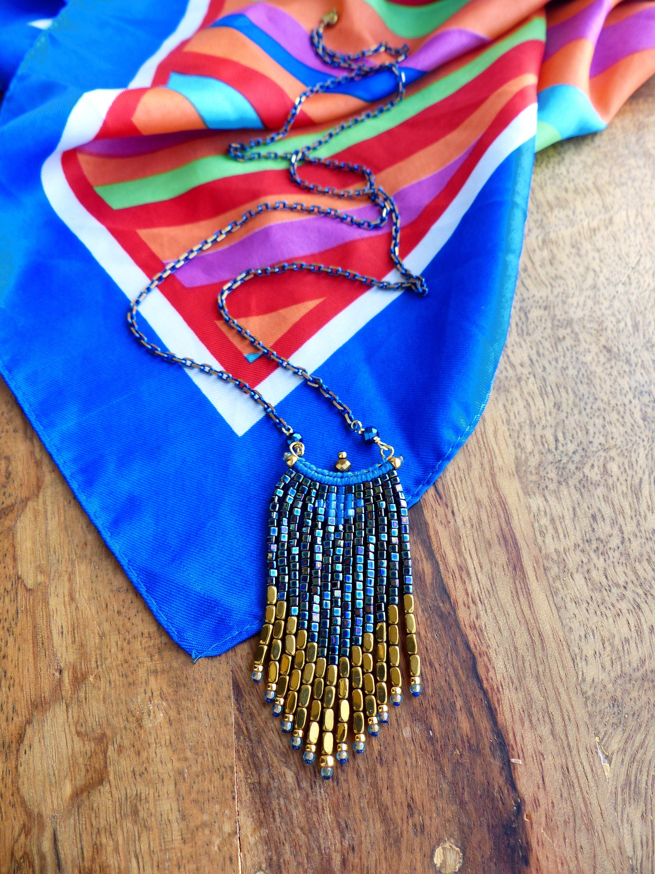 Peut inclure: Un collier de perles dorées et bleues avec un design en frange. Le collier est suspendu à une écharpe à motifs colorés.