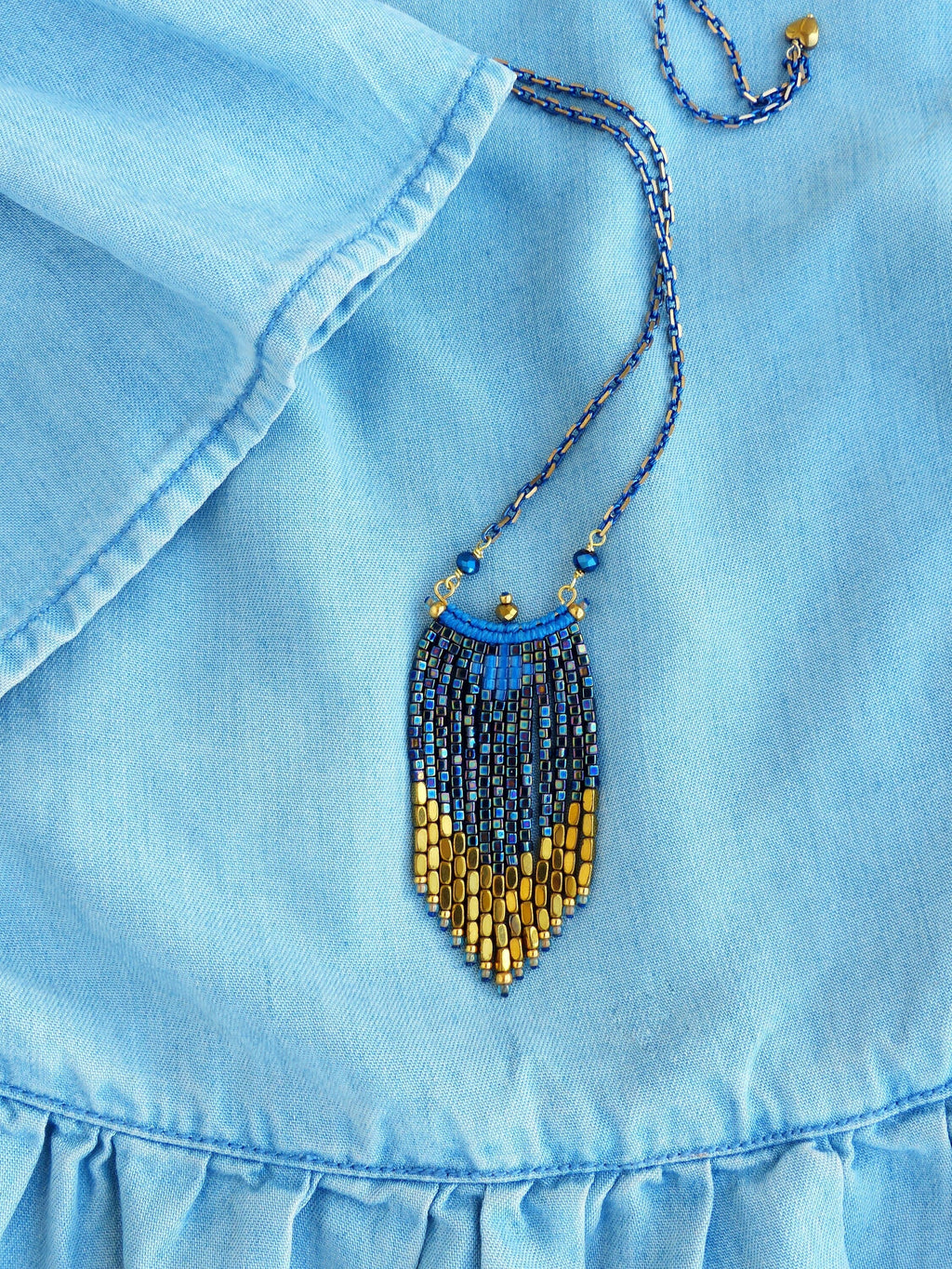 Peut inclure: Un collier de perles bleu et or avec un pendentif en frange. Le collier est sur un fond en jean bleu.