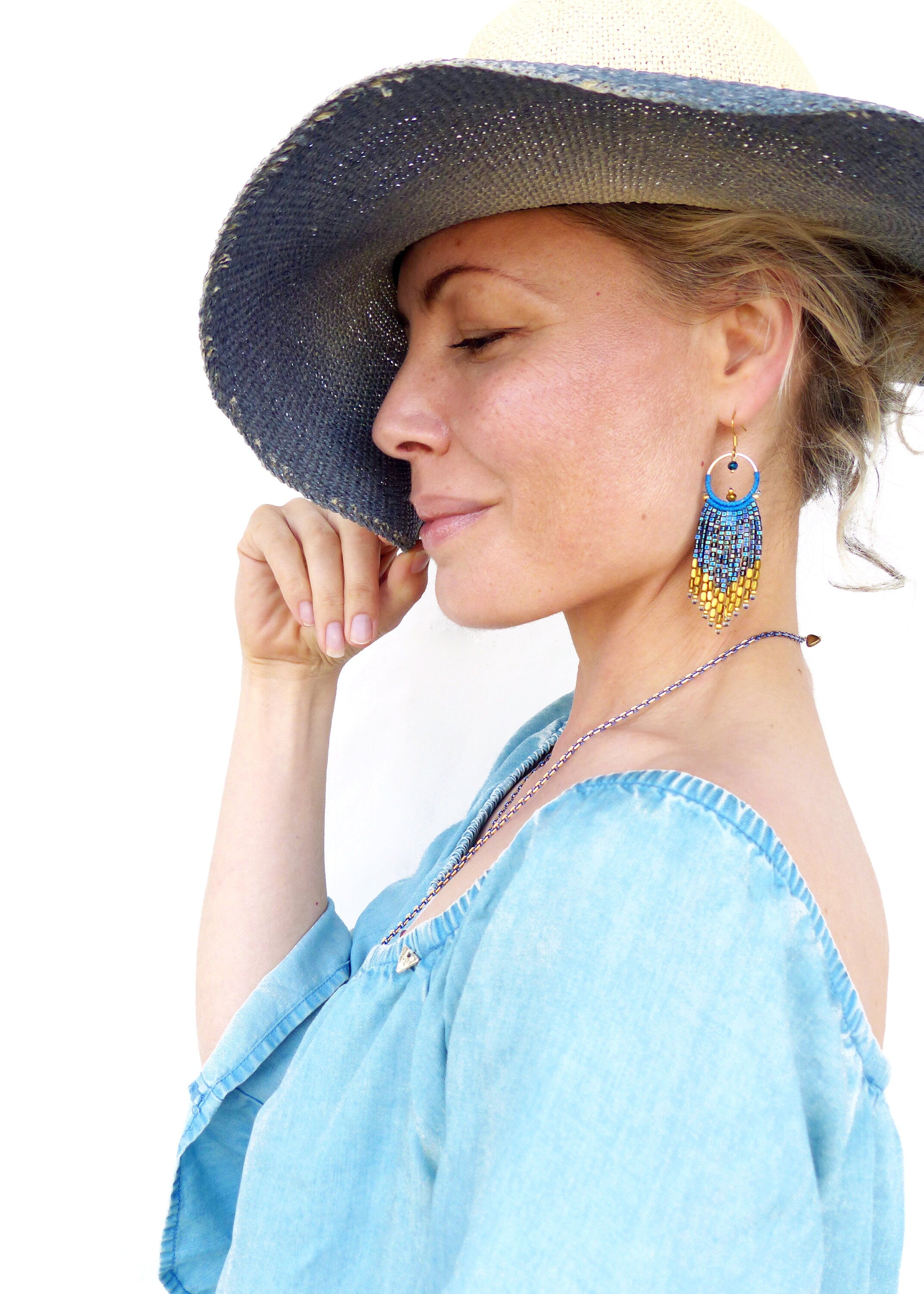 Peut inclure: Une femme porte un haut bleu en jean à épaules dénudées et un chapeau de paille à larges bords avec un bandeau bleu. Elle porte des boucles d'oreilles à franges bleues et dorées. Un collier argenté est également visible.