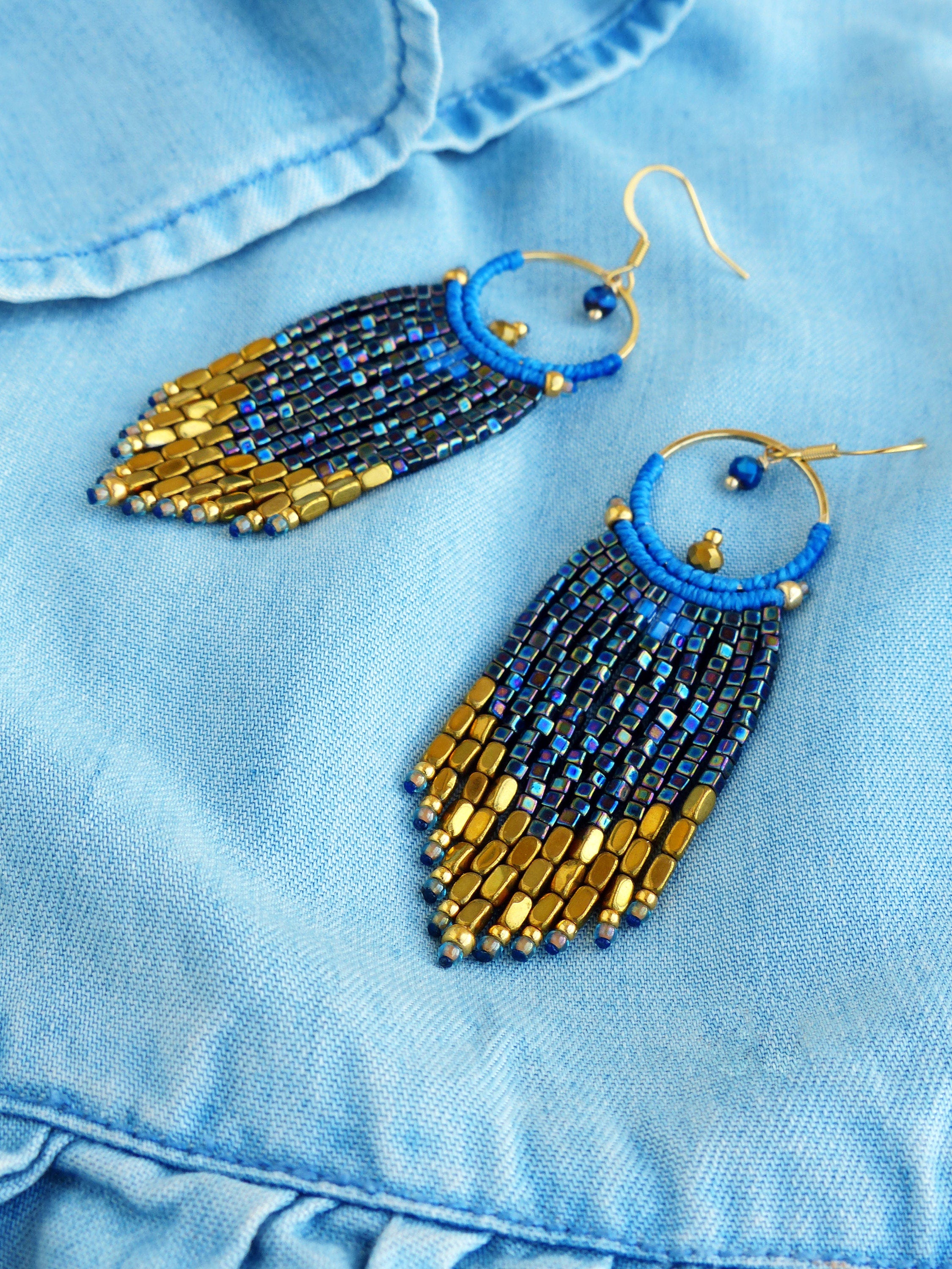 Peut inclure: Une paire de boucles d'oreilles faites à la main avec un design à franges. Les boucles d'oreilles présentent un cercle en métal doré et une frange de perles bleues qui se transforme en perles dorées en bas. Elles sont présentées sur un tissu en jean bleu clair.