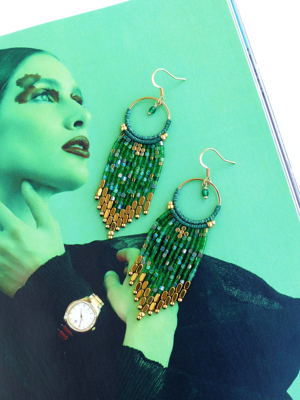 Peut inclure: Une paire de longues boucles d'oreilles en perles vertes et dorées avec un anneau en haut. Les boucles d'oreilles sont suspendues à un magazine.