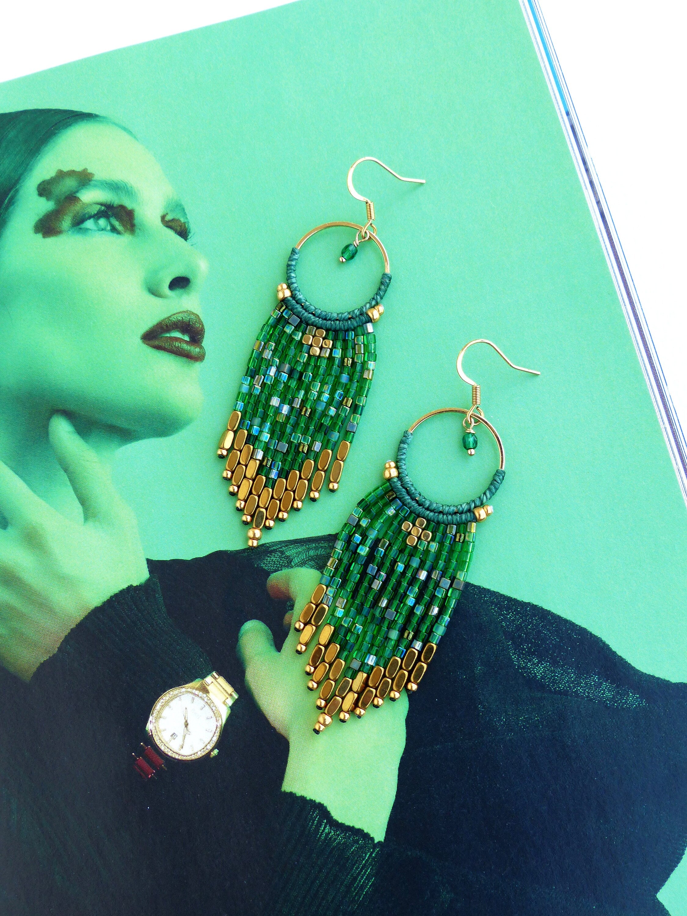 Peut inclure: Une paire de longues boucles d'oreilles en perles vertes et dorées avec un anneau en haut. Les boucles d'oreilles sont suspendues à un magazine.