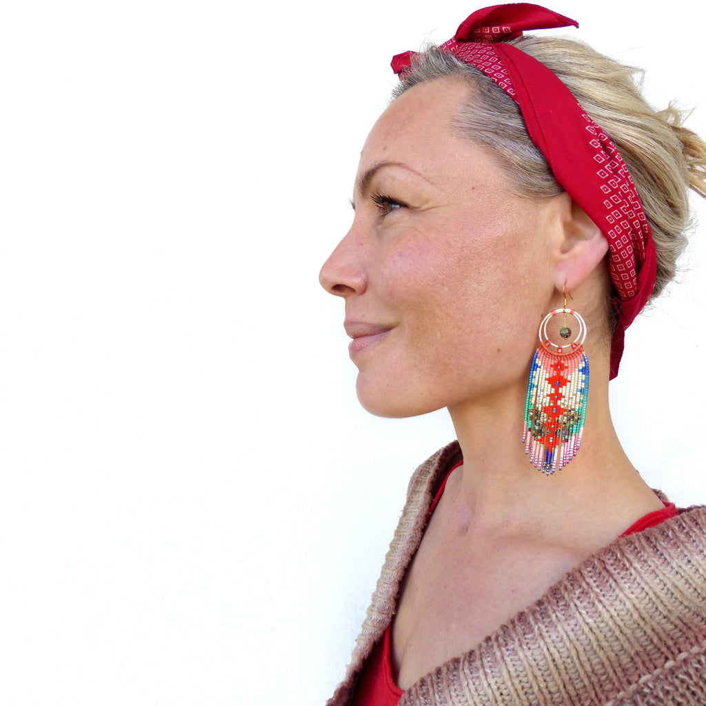 Peut inclure: Une femme portant un bandana rouge et de longues boucles d'oreilles en perles colorées. Les boucles d'oreilles présentent un motif circulaire avec une petite pierre ronde brune au centre.