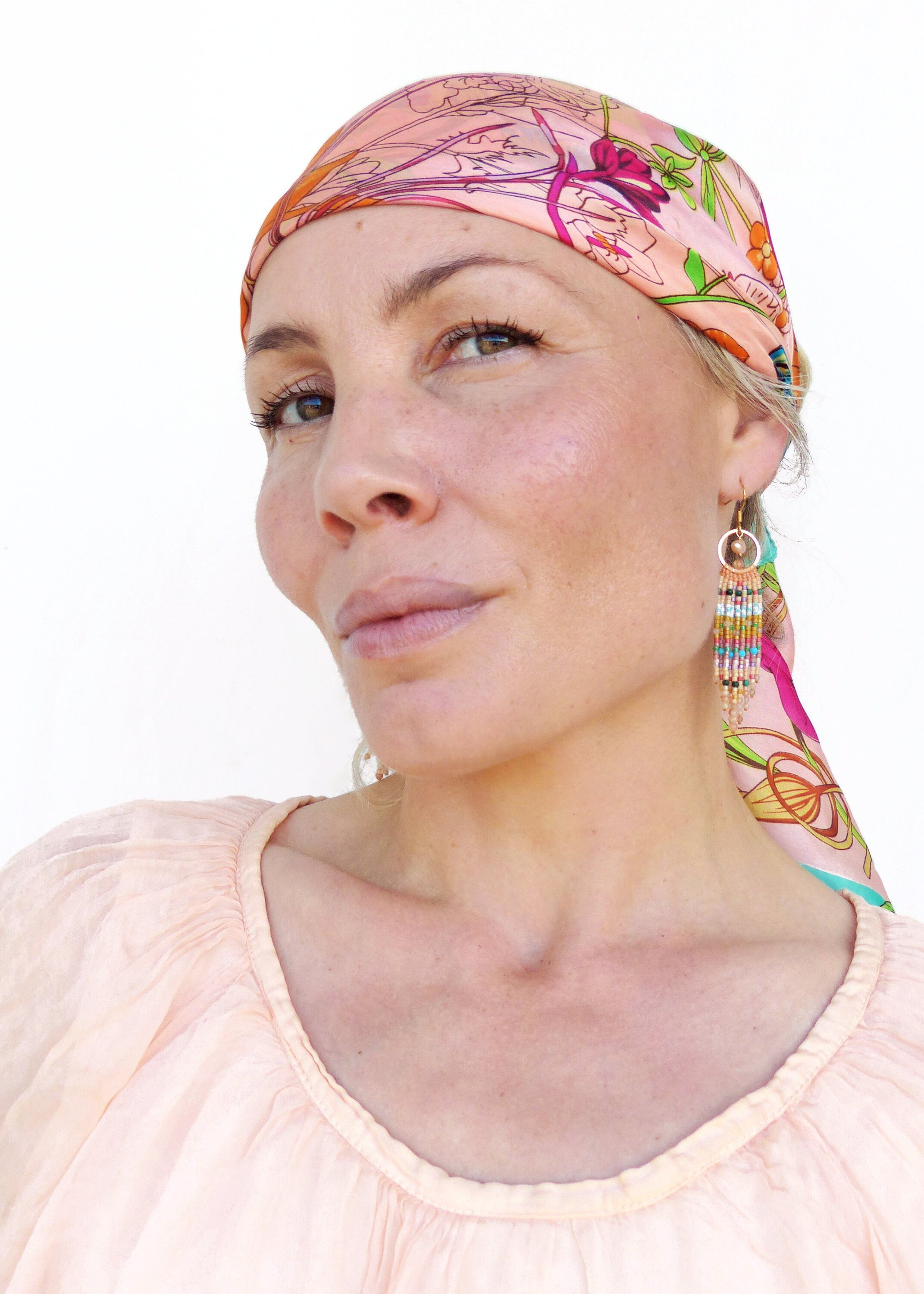 Peut inclure: Une femme aux cheveux blonds porte un foulard à motifs floraux roses et des boucles d'oreilles longues et perlées. Elle porte un chemisier rose clair.