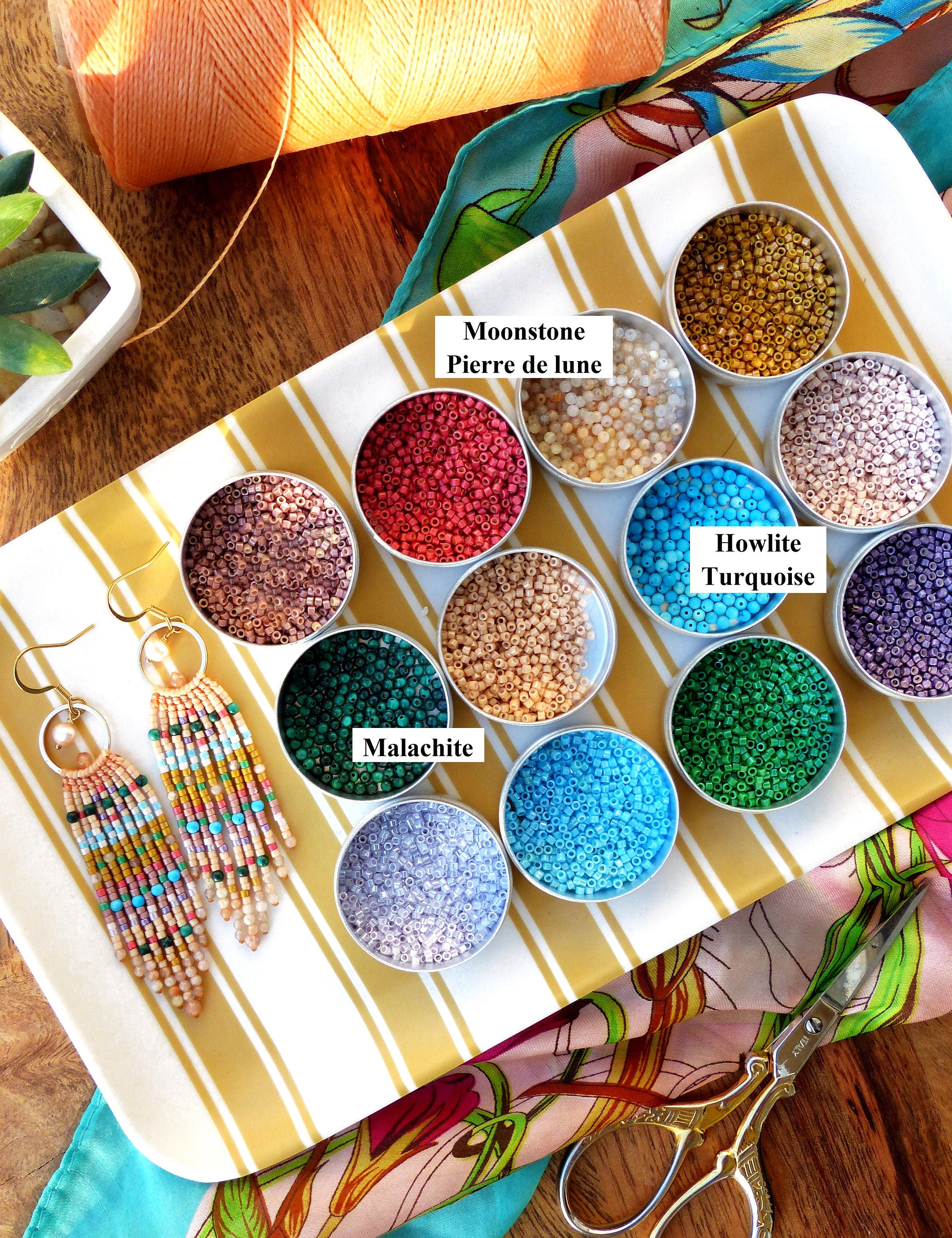 Peut inclure: Un plateau contenant de petits récipients remplis de perles colorées, étiquetés avec des noms comme "Pierre de lune", "Howlite Turquoise" et "Malachite". Deux paires de boucles d'oreilles en perles sont sur le plateau, une paire est rose, blanche et bleue, et l'autre paire est multicolore.