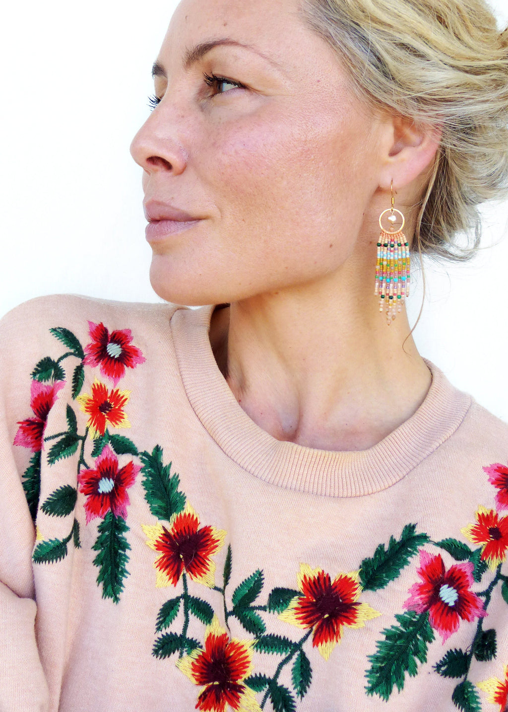 Peut inclure: Une femme portant un pull rose clair avec des fleurs brodées. Elle porte des boucles d'oreilles créoles dorées avec des franges de perles colorées.