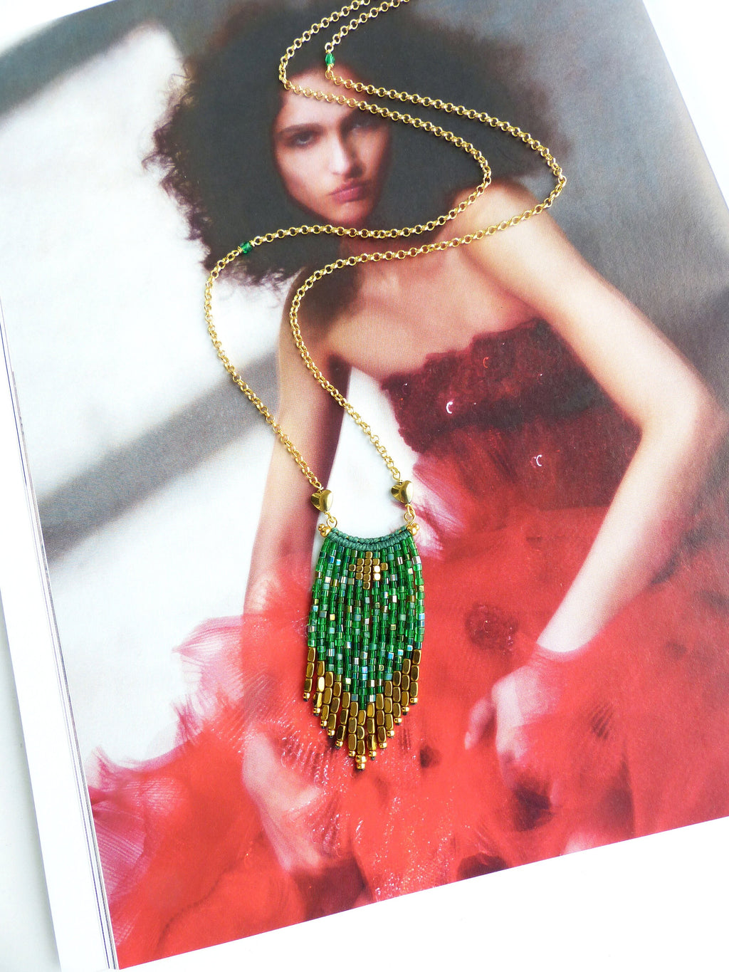Peut inclure: Un collier en chaîne dorée avec un pendentif en franges de perles vertes et dorées. Le collier est sur un magazine avec une femme portant une robe rouge.