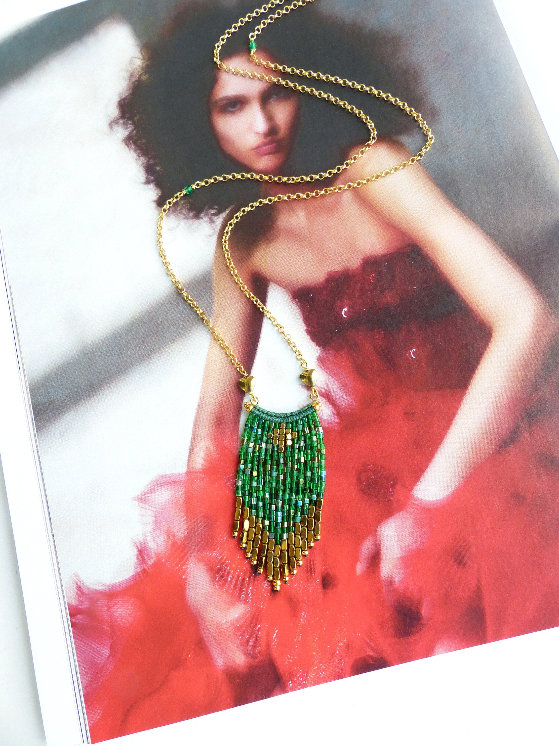 Peut inclure: Un collier en chaîne dorée avec un pendentif en franges de perles vertes et dorées. Le collier est sur un magazine avec une femme portant une robe rouge.