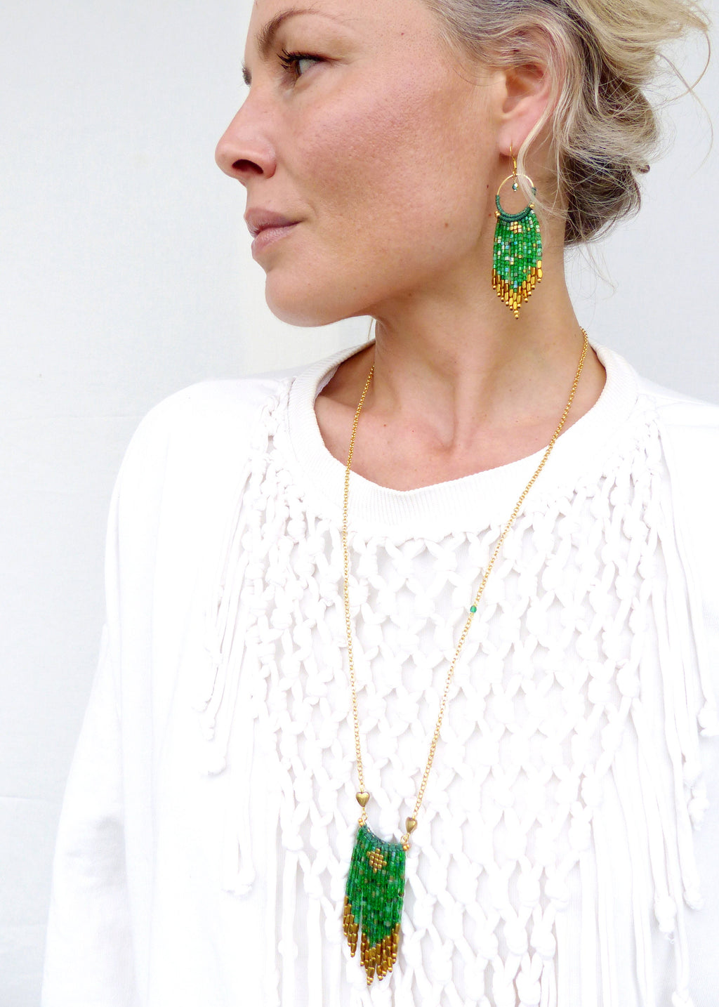 Peut inclure: Un collier en chaîne dorée avec un pendentif en perles vertes et dorées pend autour du cou d'une personne portant un haut en crochet blanc. Le pendentif est en forme de larme avec un motif géométrique. La personne porte également des boucles d'oreilles assorties en perles vertes et dorées.
