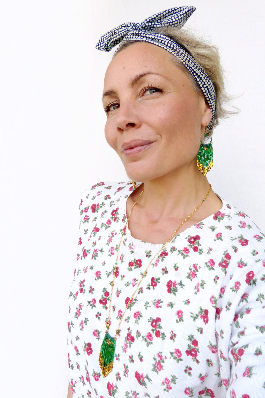 Peut inclure: Une femme portant une chemise à fleurs blanches avec un collier en chaîne dorée et des boucles d'oreilles en perles vertes et dorées. Elle porte également un bandeau à pois noirs et blancs.