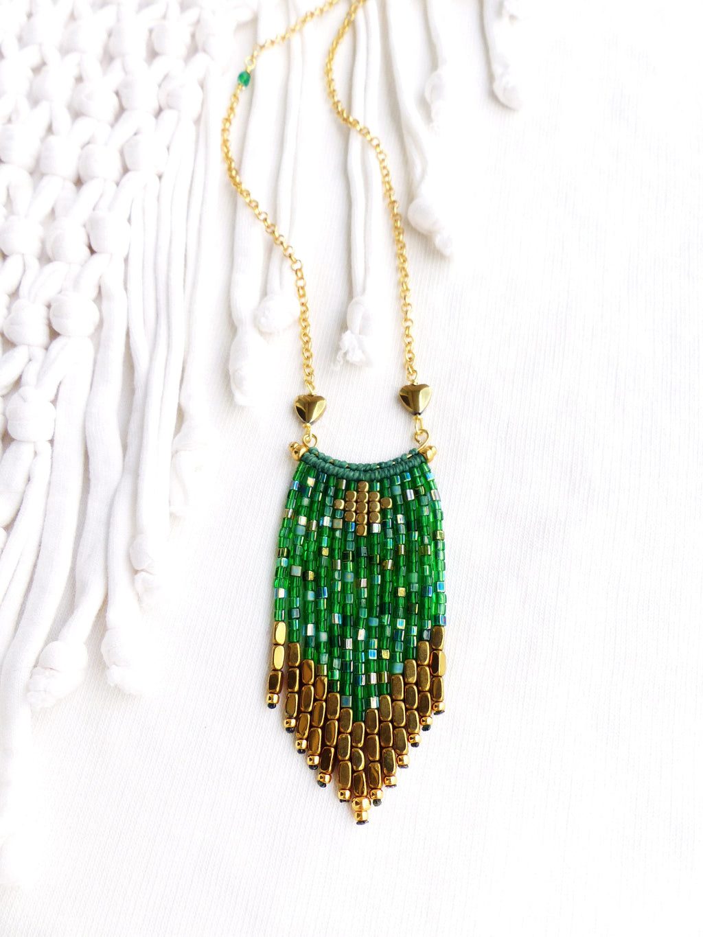 Peut inclure: Un collier en chaîne dorée avec un pendentif en franges de perles vertes et dorées. Le pendentif est composé de petites perles carrées dans des tons de vert et d'or, avec une perle en forme de cœur dorée en haut.