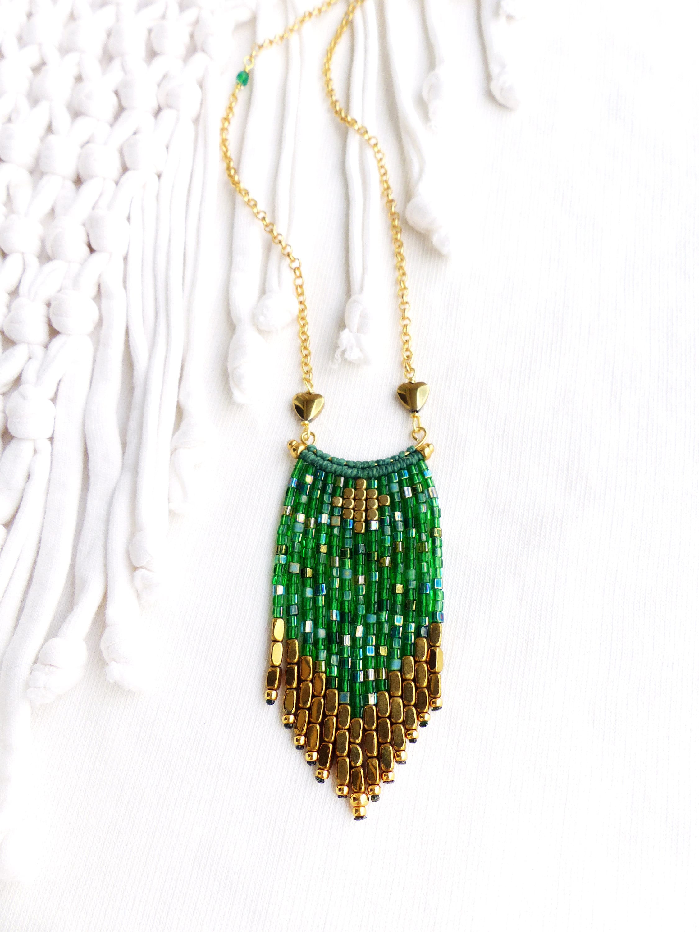 Peut inclure: Un collier en chaîne dorée avec un pendentif en franges de perles vertes et dorées. Le pendentif est composé de petites perles carrées dans des tons de vert et d'or, avec une perle en forme de cœur dorée en haut.